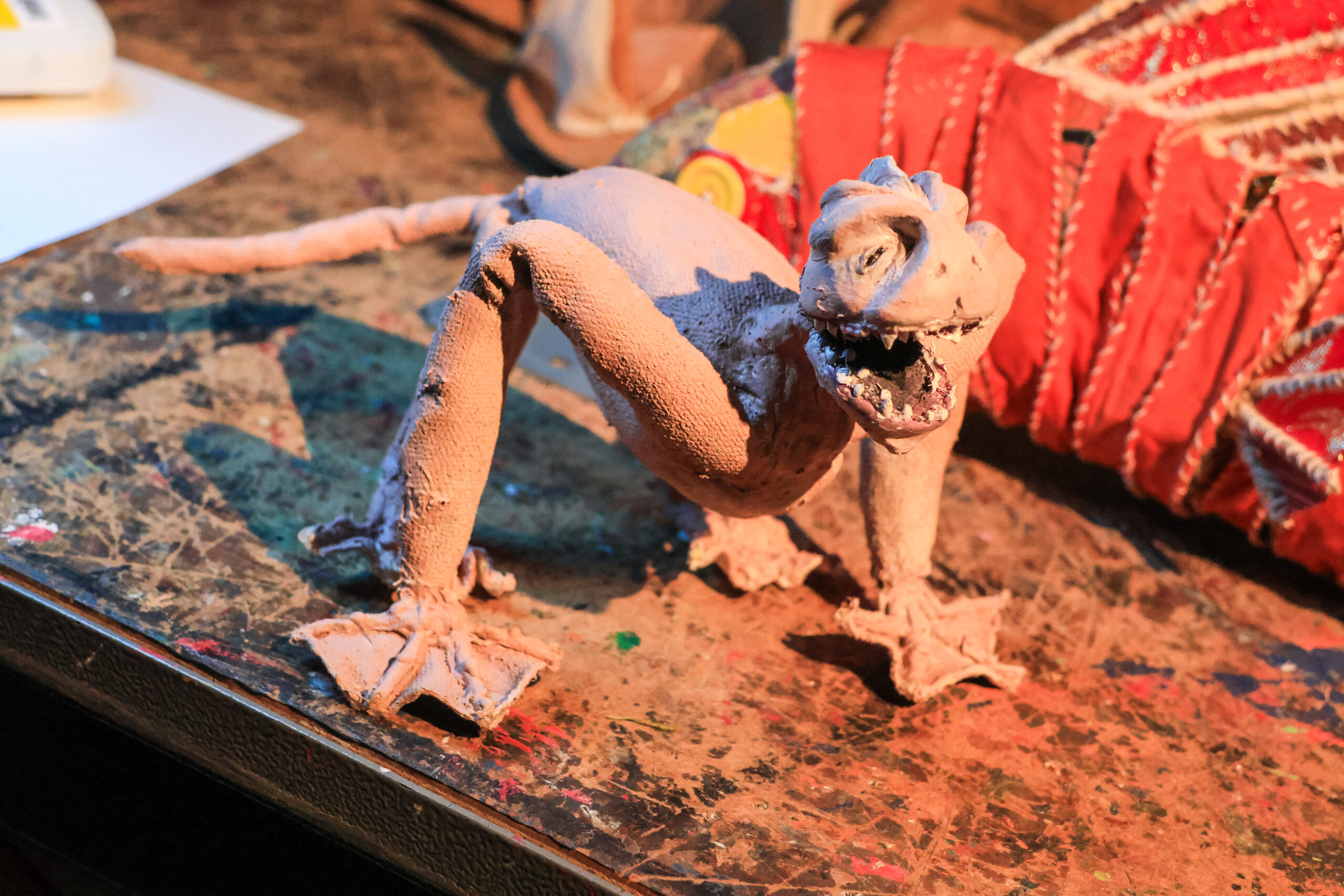 A handmade clay sculpture of a small, humanoid creature resembling a lizard. It has a wide mouth with sharp teeth and a textured body, supported by two legs and a long tail. The figure is positioned on a colorful, paint-splattered surface, alongside a red decorated item in the background. The lighting casts shadows that emphasize the creature's features.