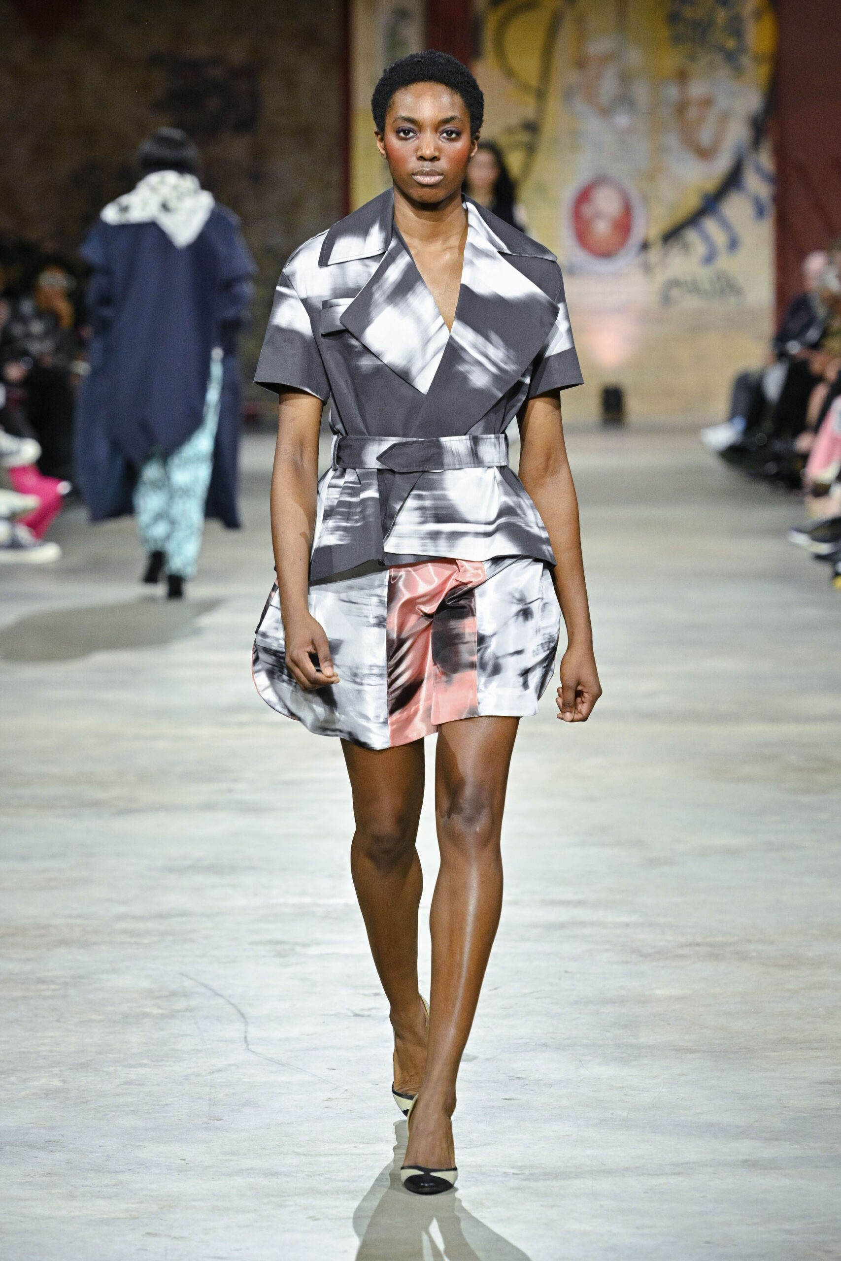 A model walks down a runway wearing a stylish ensemble featuring a gray dress with abstract black and white patterns and a pink and gray skirt. The outfit is complemented by black and white shoes. The setting showcases an audience seated in the background, with a mix of blurred figures and a textured wall behind the model.