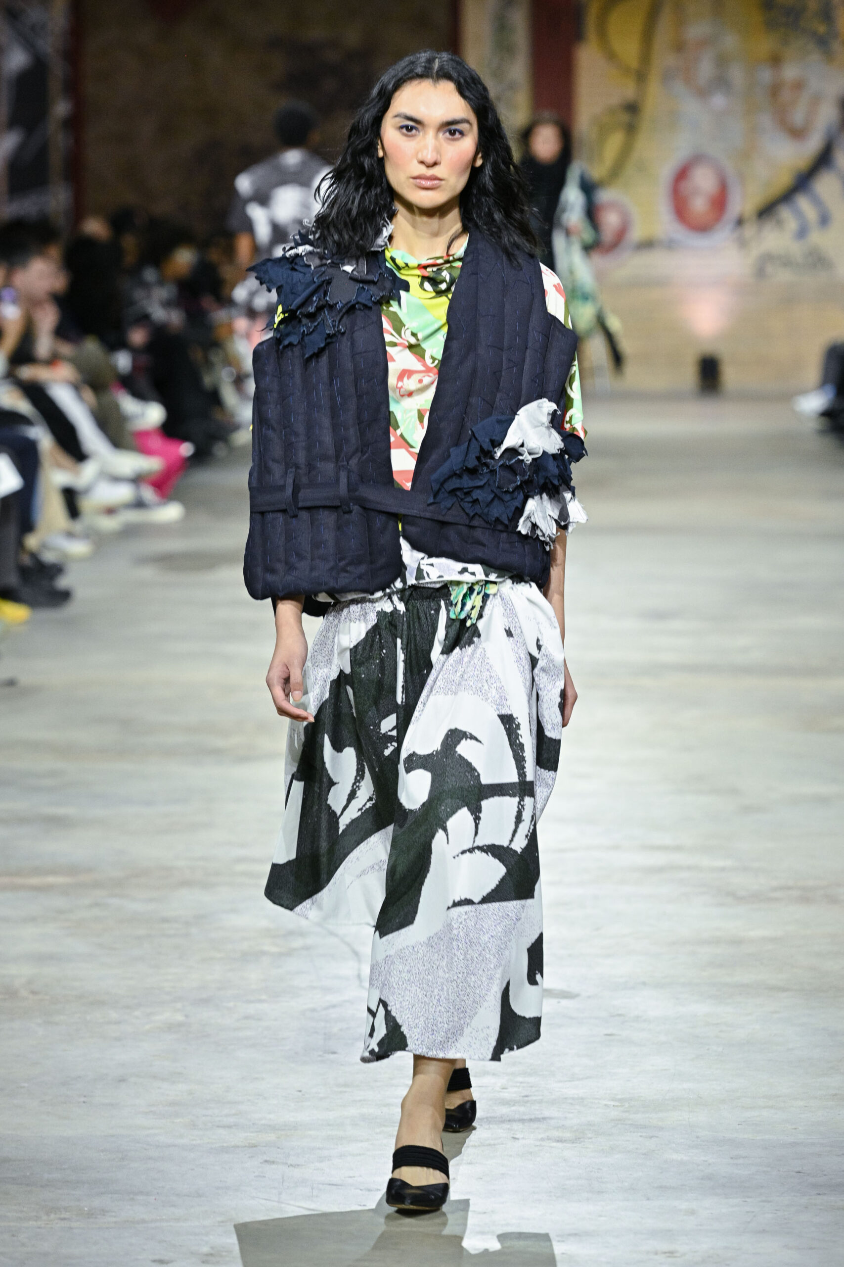 A model walks down a runway, wearing a vibrant floral top and a dark quilted vest adorned with textured fabric details. She pairs it with a flowing, patterned skirt featuring black and white designs. The setting has an artistic backdrop with an audience visible in the background, observing the fashion show. The model wears black shoes and has wavy, shoulder-length hair.