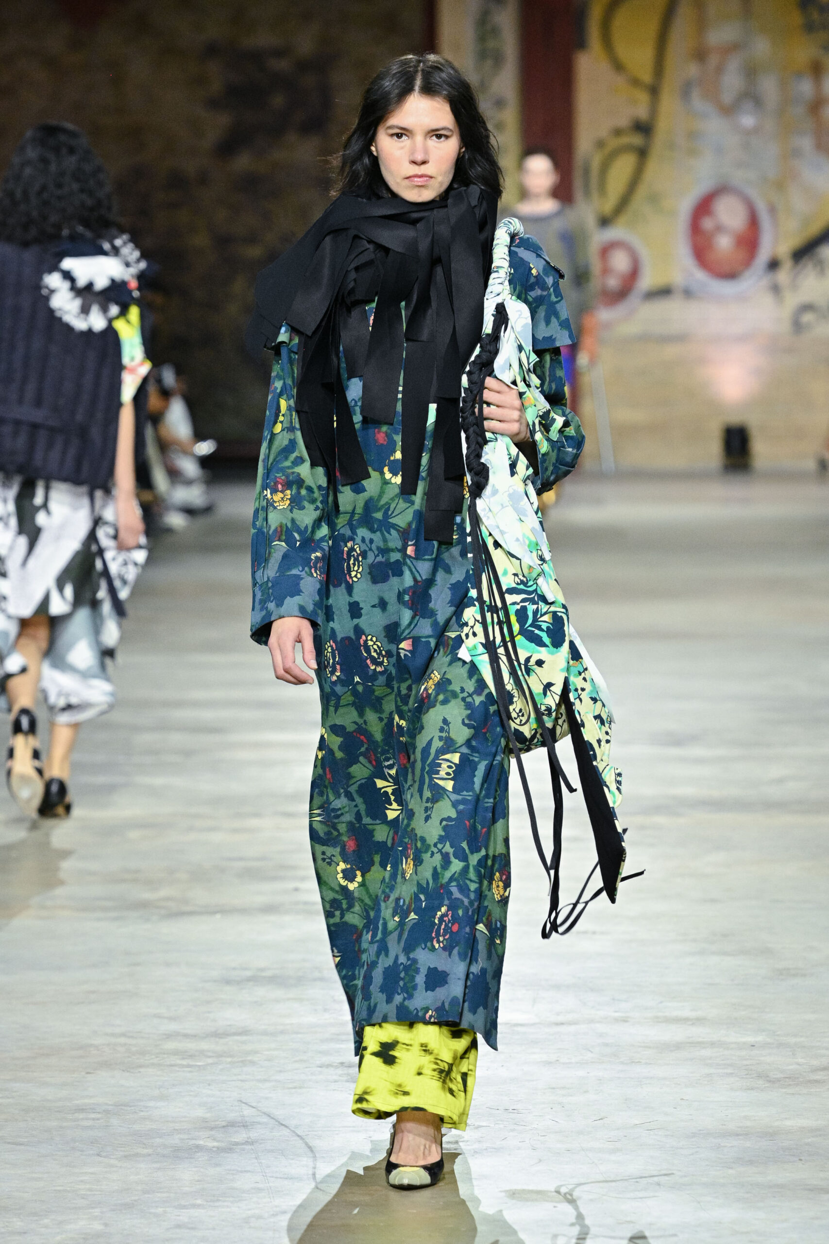 A model walks down a runway, wearing a long, floral dress in shades of blue and green, layered over a bright yellow garment at the bottom. The outfit is accessorized with a large black scarf and a patterned bag slung over one shoulder. The runway features a spacious and industrial setting, with other models in the background.
