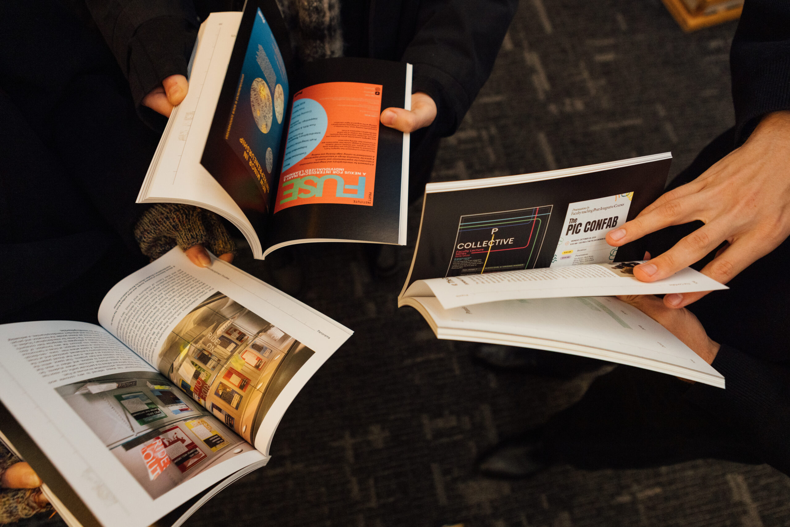 Several hands are holding open books, displaying colorful pages and graphics. The books feature various designs, layouts, and text, with one showing images of framed illustrations on a wall. The scene captures a collaborative moment as individuals explore the contents together, set against a dark carpeted surface.