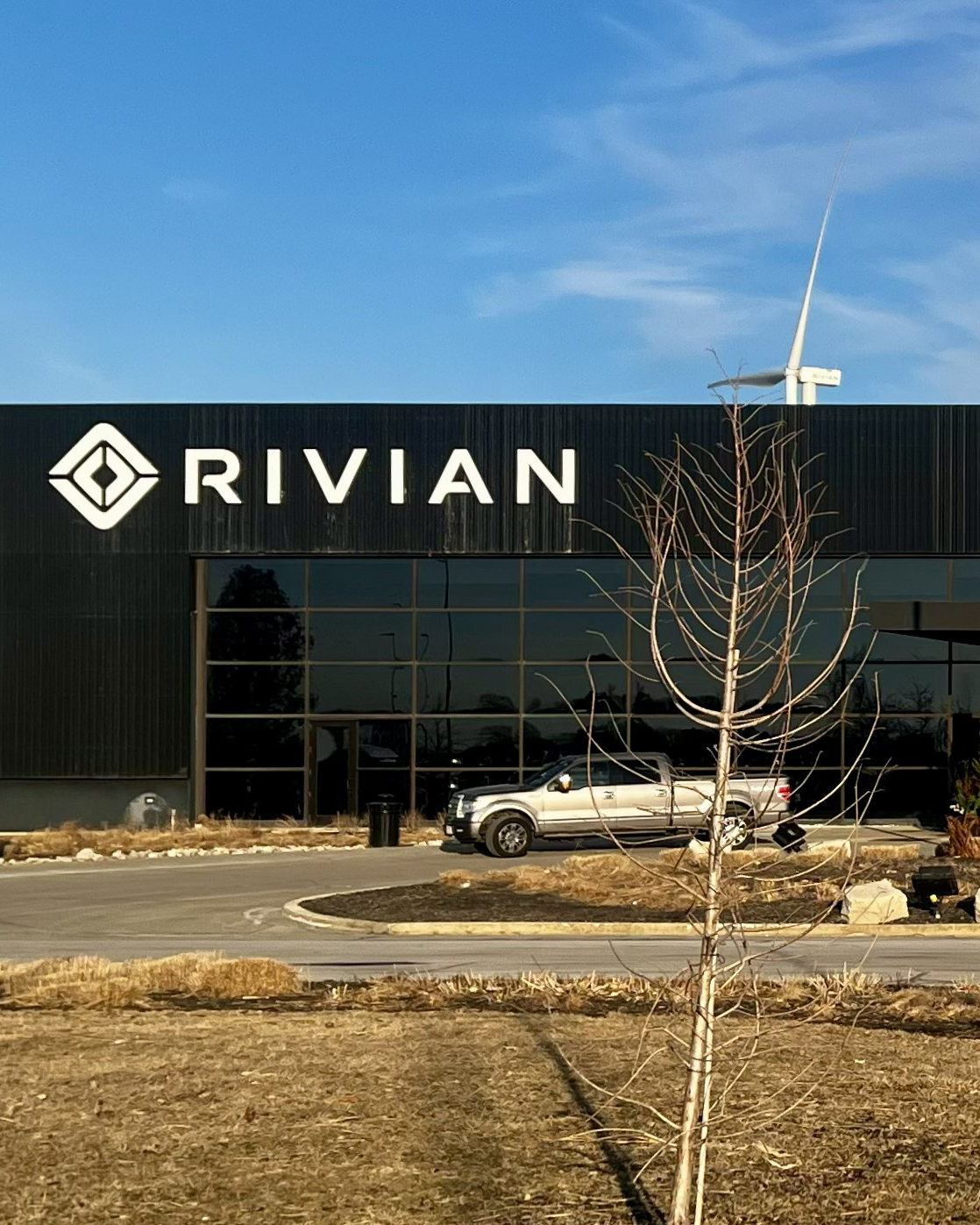 A building with a dark exterior displays the logo and name "RIVIAN" prominently on its facade. In front of the building, a bare tree stands, and a silver vehicle is parked nearby. The sky above is clear with a few clouds visible. The area around the building is landscaped with dry grass and rocks.