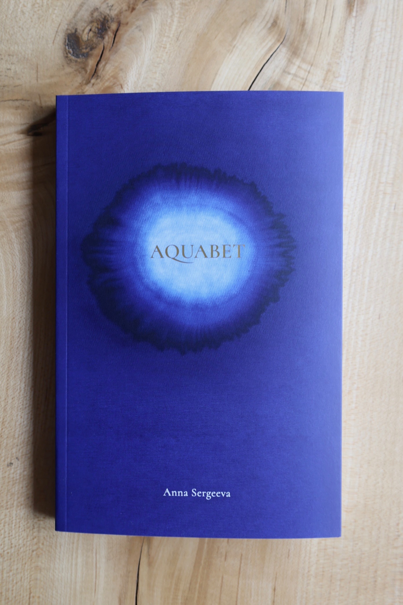 Cover of Aquabet by Anna Sergeeva. On a deep blue background, the gold title is encircled by rings of pale blue, medium blue, and dark blue, like ink into which water has been dripped.
