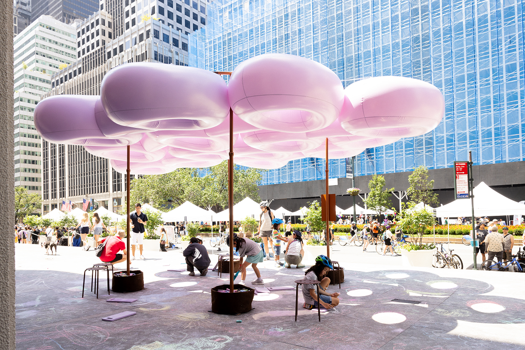 A vibrant outdoor scene featuring a large, pink, cloud-like installation supported by poles. Beneath the structure, several children and adults are engaged in activities, with some sitting on small stools and drawing on the ground with chalk. In the background, tents are set up for an event, surrounded by greenery and pedestrians, while tall buildings and city life are visible.