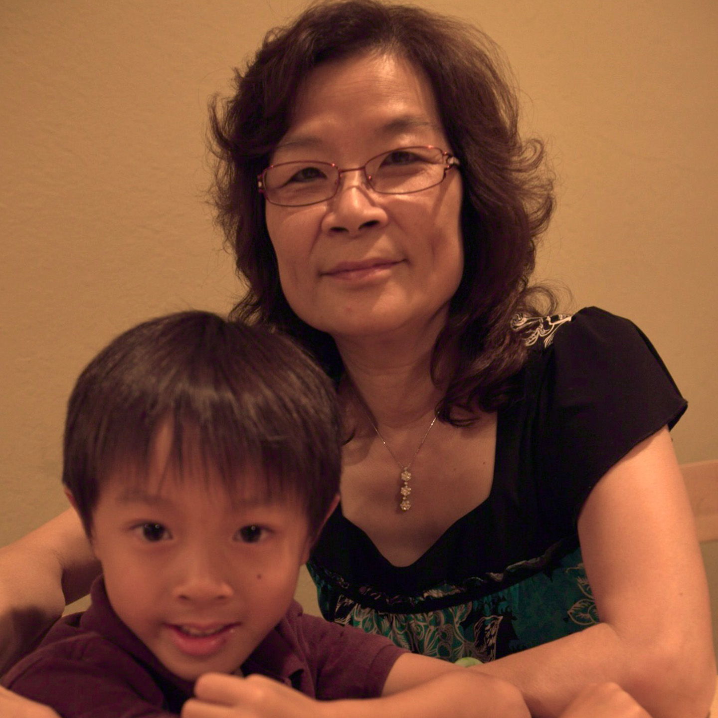 A woman with wavy dark hair wearing glasses sits next to a young boy with short dark hair. The woman is wearing a black top with colorful patterns. They both smile at the camera, with the boy in front and looking slightly to the side. The background is a plain beige wall.