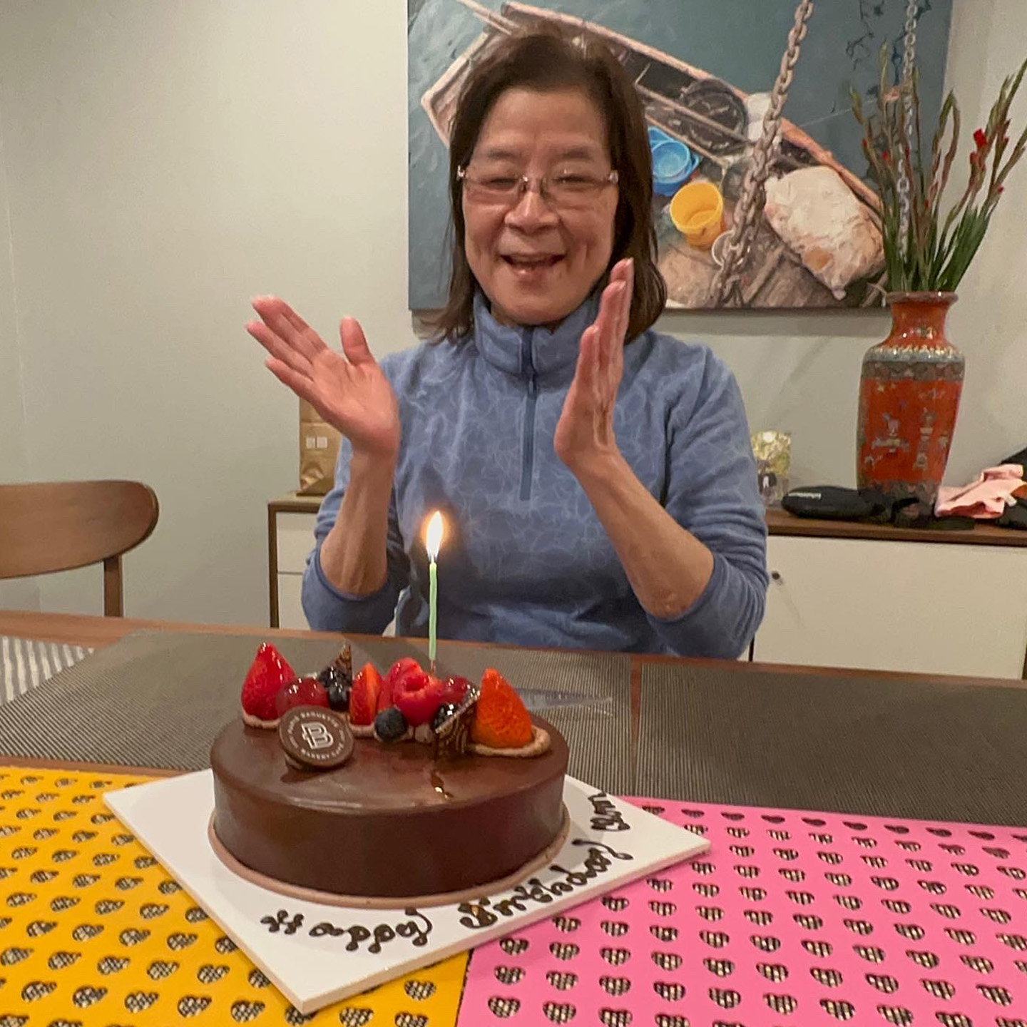 A woman is sitting at a table with a birthday cake in front of her, adorned with various fruits and a single green candle lit on top. She is smiling and clapping her hands in celebration. The background features a colorful art piece and a decorative vase. The table is covered with a patterned tablecloth in pink and yellow. The cake has "Happy Birthday" written in icing.