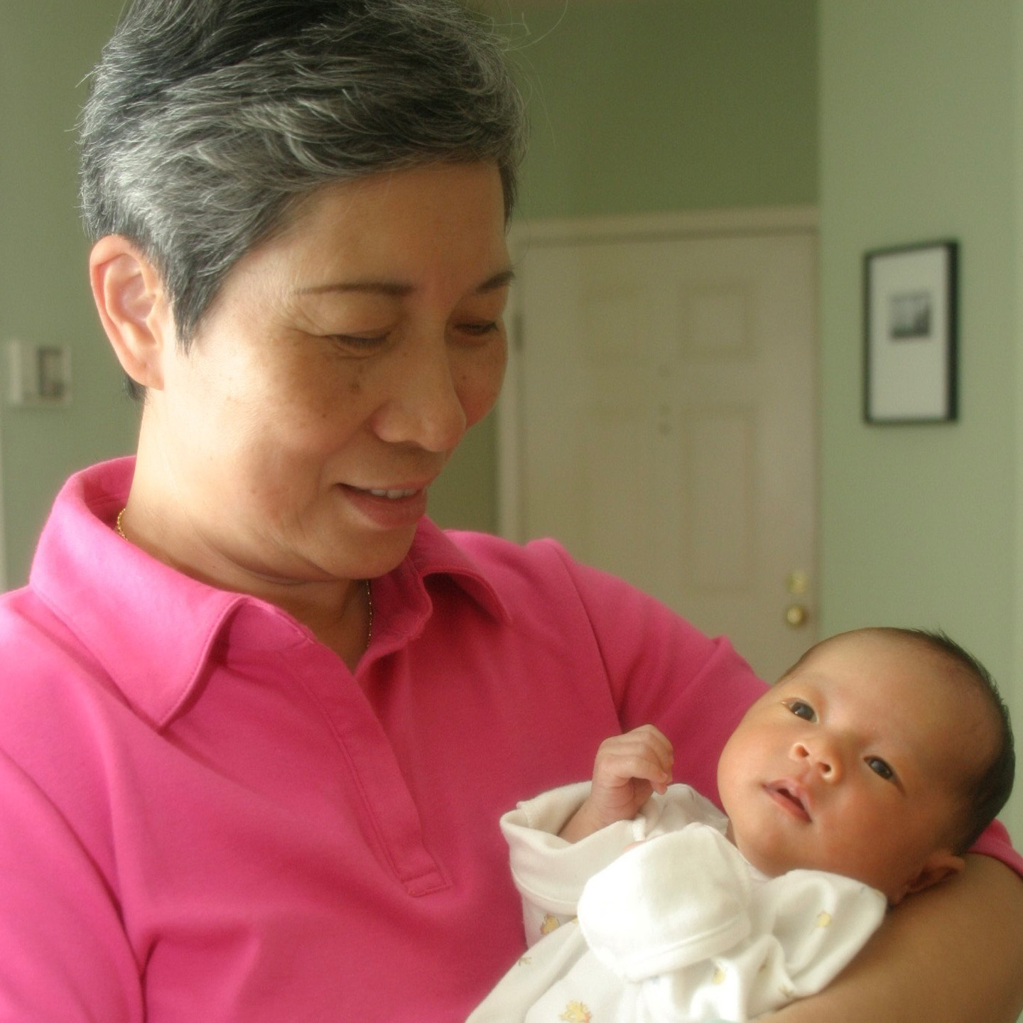 A smiling woman with short gray hair is holding a small baby in a white outfit. The woman is wearing a pink polo shirt and looking down at the baby, who is gazing back at her with curiosity. The background features a softly lit interior with a door and a framed picture on the wall.