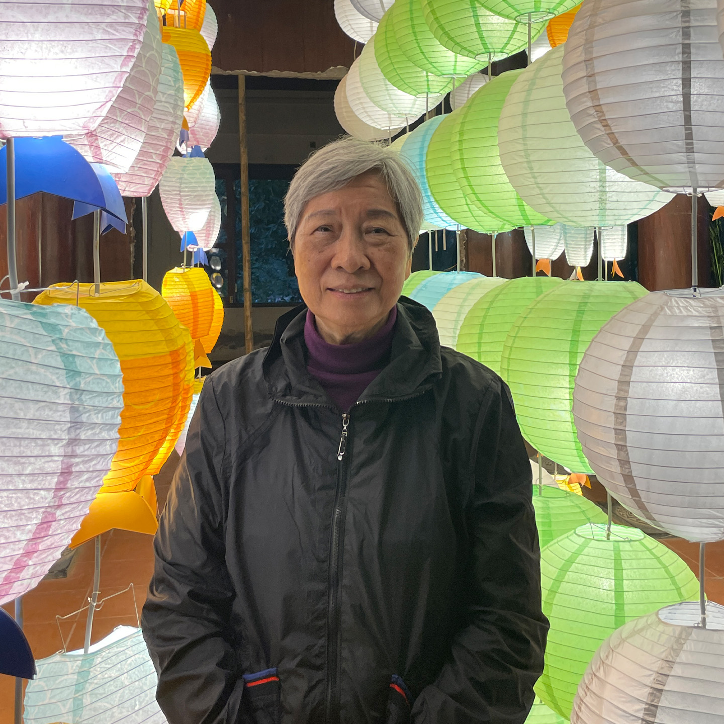 An older adult with short gray hair is standing in front of a colorful display of paper lanterns. The lanterns are in various hues, including orange, green, white, and blue, hanging above him and behind him. He is wearing a black jacket and purple turtleneck, smiling gently at the camera. The setting appears to be indoor, illuminated by the soft glow of the lanterns.