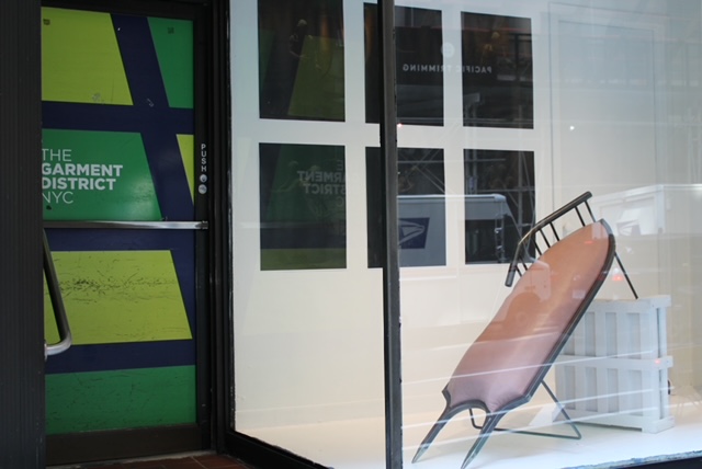 A storefront displays a glass window with a folding lounge chair placed inside on a white platform. Behind the chair, there are several framed artworks or posters with dark backgrounds. The entrance door features colorful graphics and the text "THE GARMENT DISTRICT NYC." The overall scene is clean and modern, emphasizing the unique display inside.