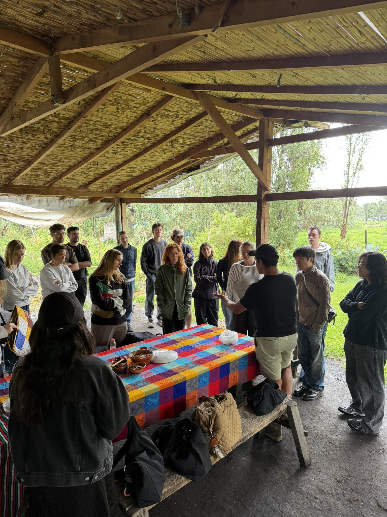 A group of people is gathered under a thatched-roof structure in an outdoor setting. They are listening attentively to a speaker, who is gesturing while standing near a colorful table covered with plates and bowls. The audience consists of diverse individuals, some wearing casual clothing, and there are bags on the ground. Lush greenery is visible in the background, suggesting a rural environment.