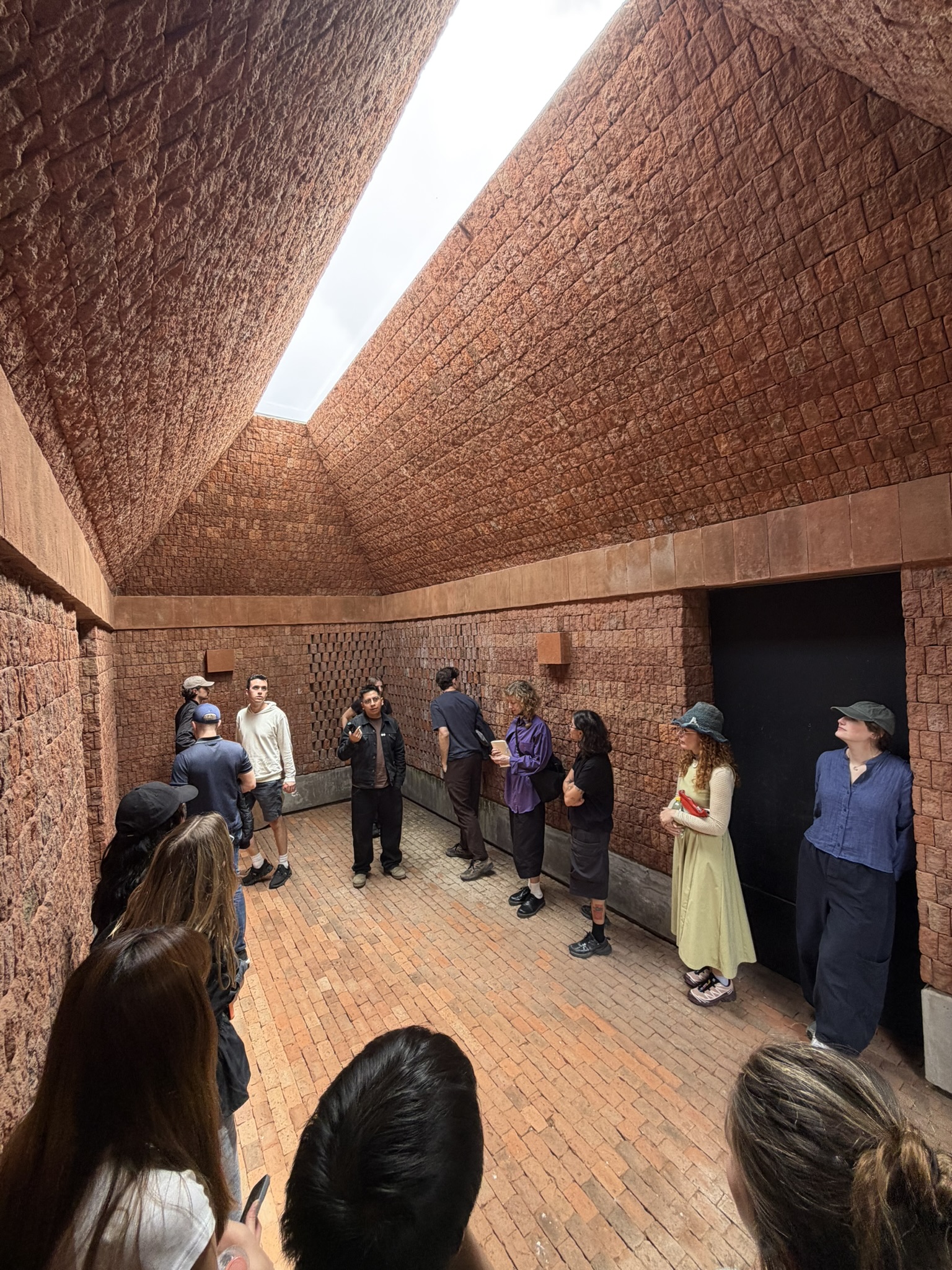A group of people gathered in a brick-walled room with a slanted ceiling, illuminated by natural light coming from an opening at the top. One person, standing in the center, appears to be speaking or presenting, while others listen attentively, some leaning against the walls. The individuals are dressed in casual clothing, and the atmosphere seems to be one of discussion or learning.