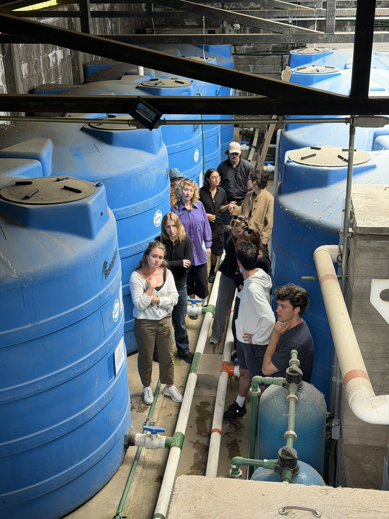 A group of people stands among large blue water tanks in a warehouse setting. The individuals, both men and women, appear to be engaged in conversation while observing their surroundings. Some are dressed casually, and there's a mix of hair colors and styles. The environment features visible pipes and machinery, indicating an industrial or educational setting focused on water management.
