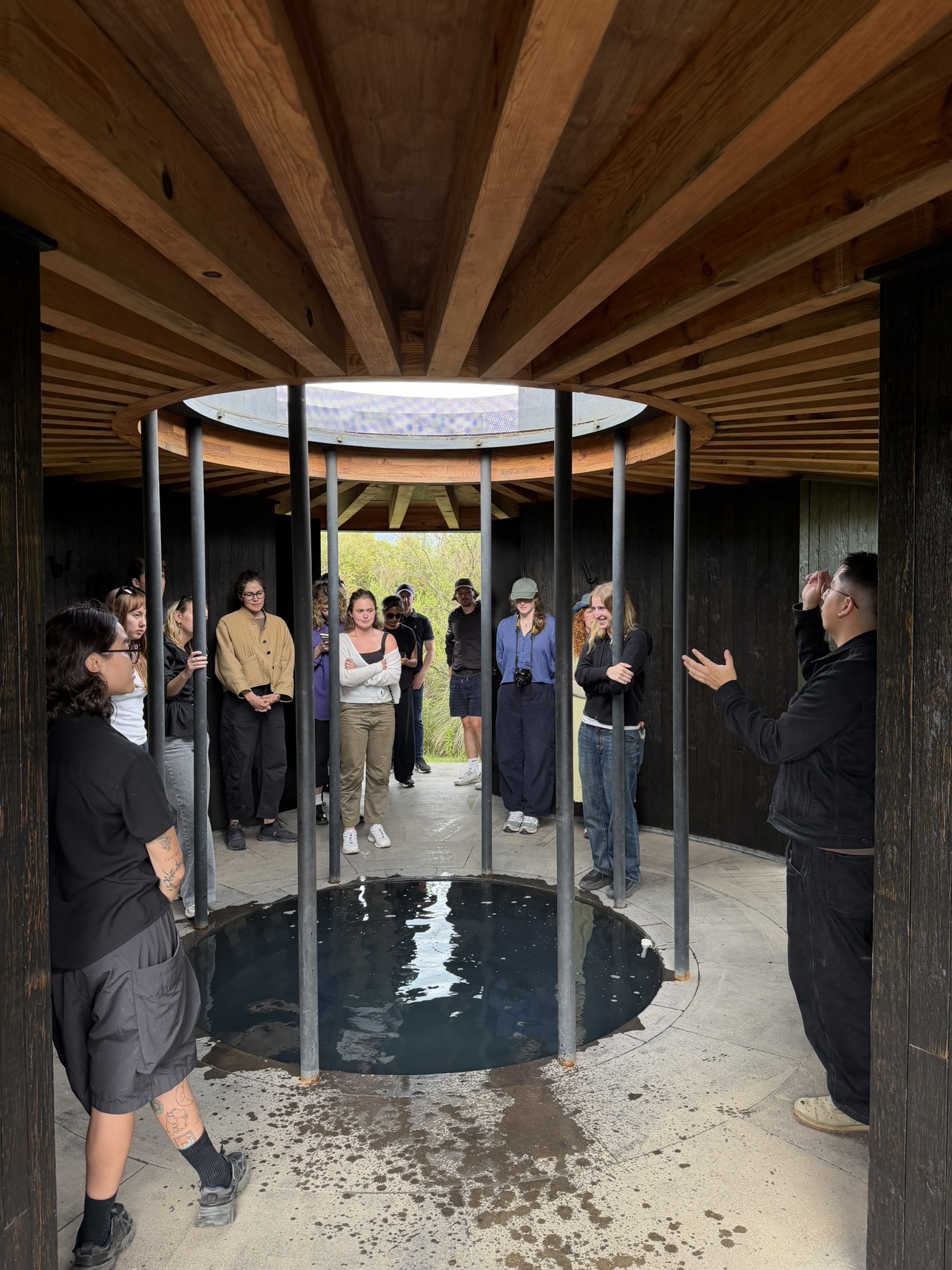 A group of diverse individuals stands inside a circular structure with wooden beams and a skylight overhead. They are gathered around a circular pool of still water at the center. One person is gesturing while speaking to the group. The atmosphere is informal and educational, with people wearing casual clothing.