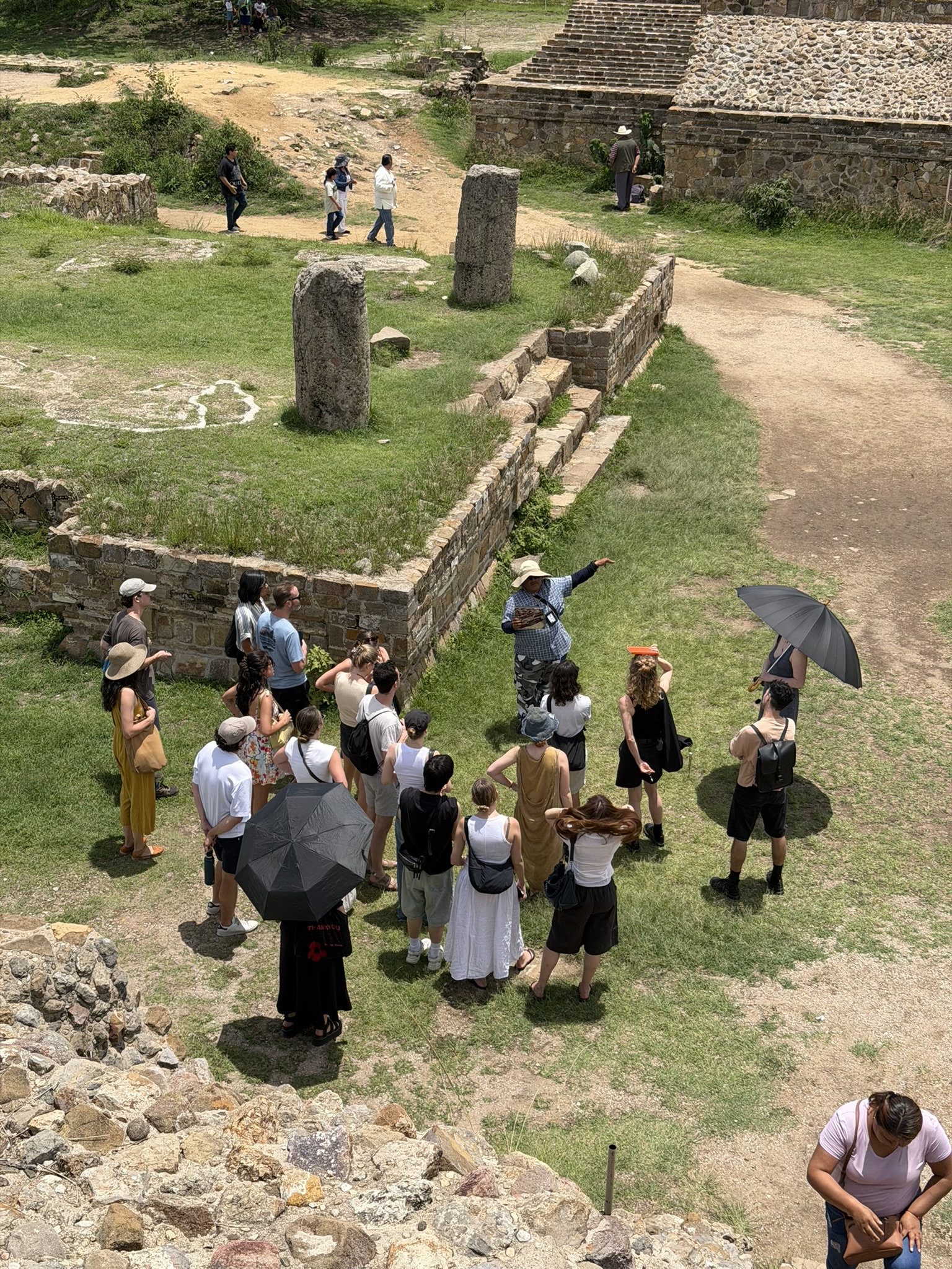 A guided tour is taking place at an archaeological site. A group of people, mostly wearing light clothing and hats for sun protection, gathers around a guide who is pointing and explaining something. Some attendees are holding umbrellas for shade. In the background, there are rocky structures and greenery. Various individuals are seen walking and exploring the area.