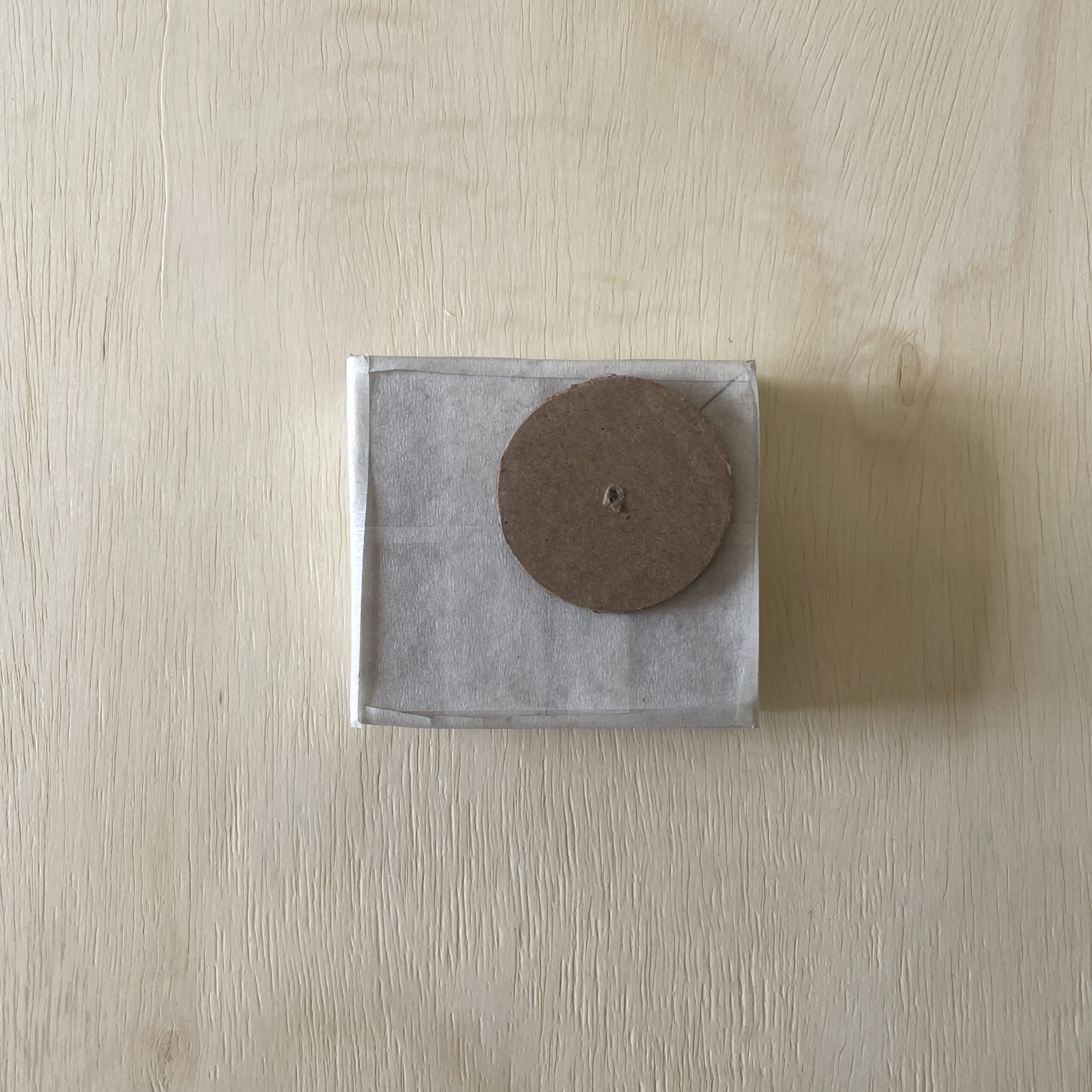 A small, square package wrapped in light brown paper is placed on a light wooden surface. On top of the package, there is a circular disk made of brown cardboard. The disk has a small hole in the center, while the edges are slightly uneven.