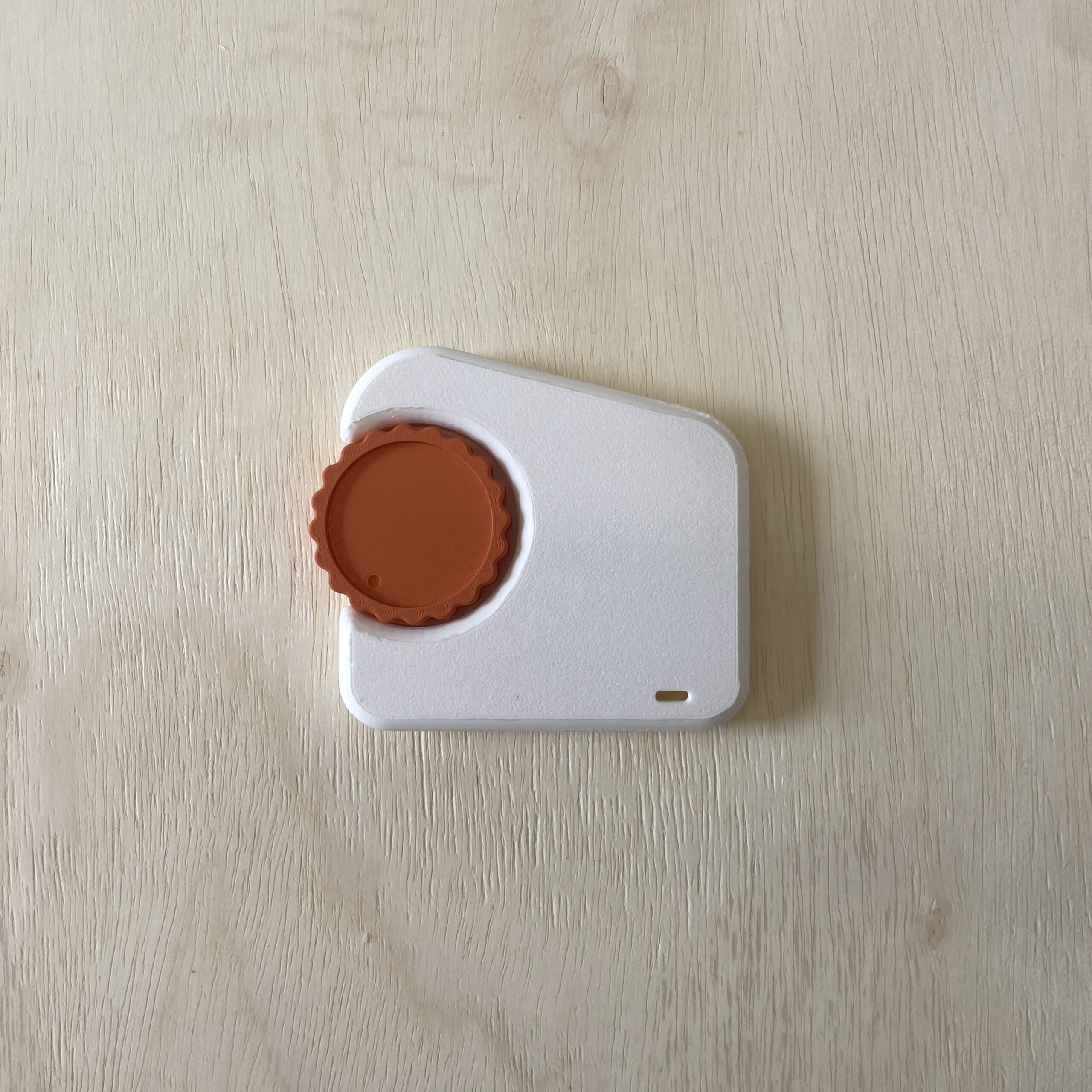 A close-up view of a small, white electronic device with a round orange button on top. The device has a slightly angled shape and a small indicator light at the bottom. It is placed on a light wooden surface with a textured finish.