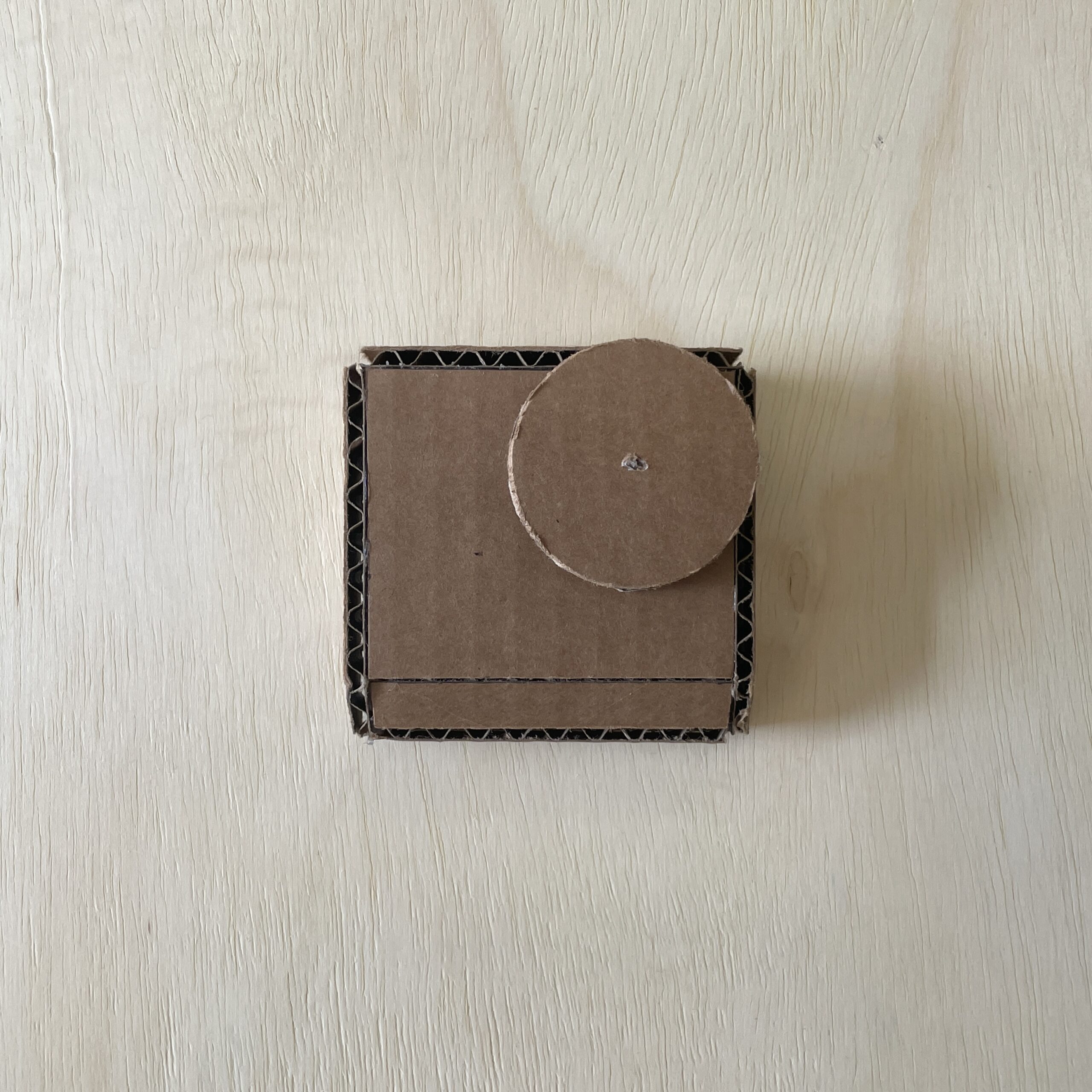 A small square cardboard box with a circular lid is placed on a light wood surface. The box has a simple, unadorned design and features a layered texture. The circular lid sits slightly off-center on top of the box. The overall appearance is minimalistic and utilitarian.