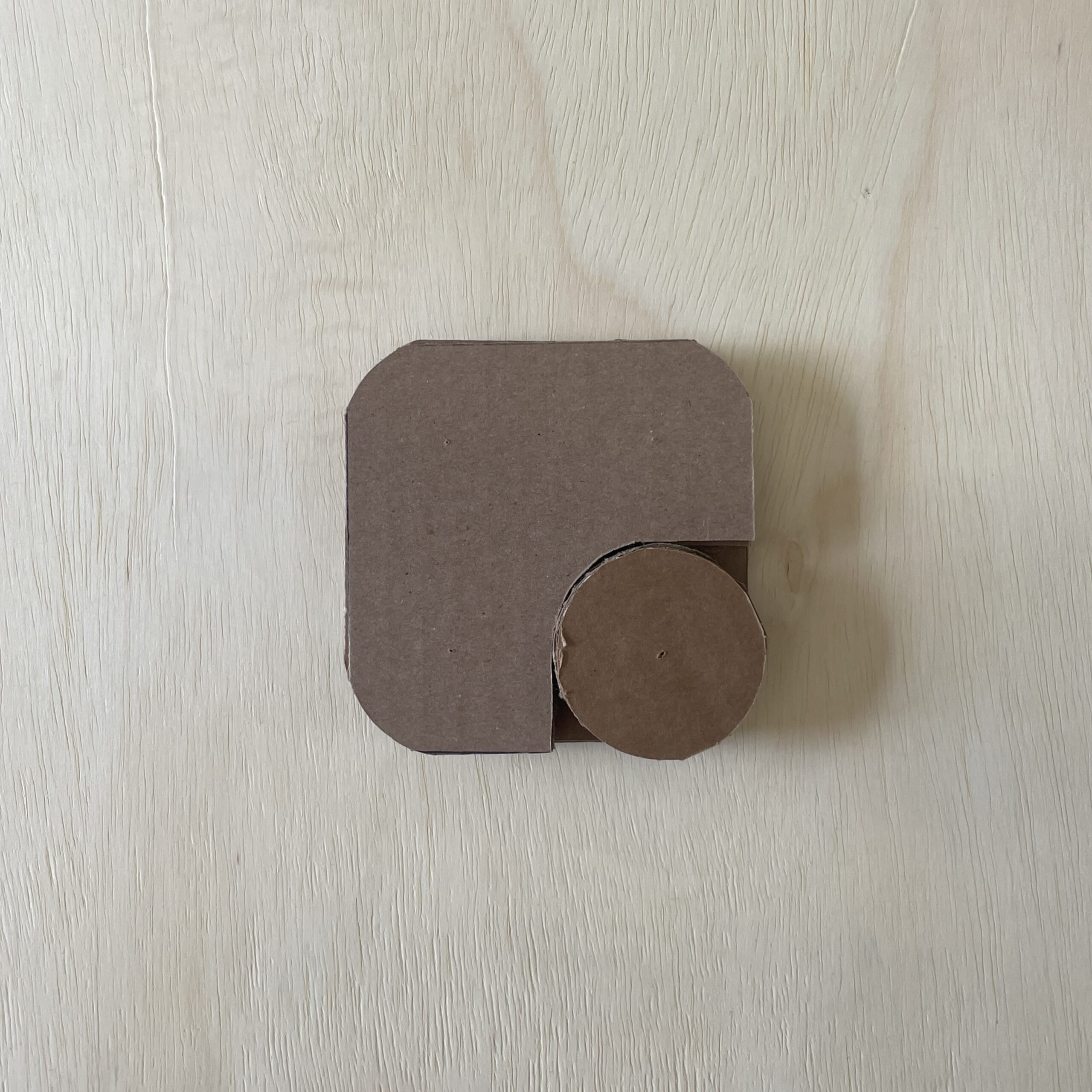A small piece of brown cardboard is displayed on a light wooden surface. The cardboard has a rounded square shape with one circular cutout on its side, resembling a simplistic design. The texture of the cardboard is visible, indicating its thickness and material.