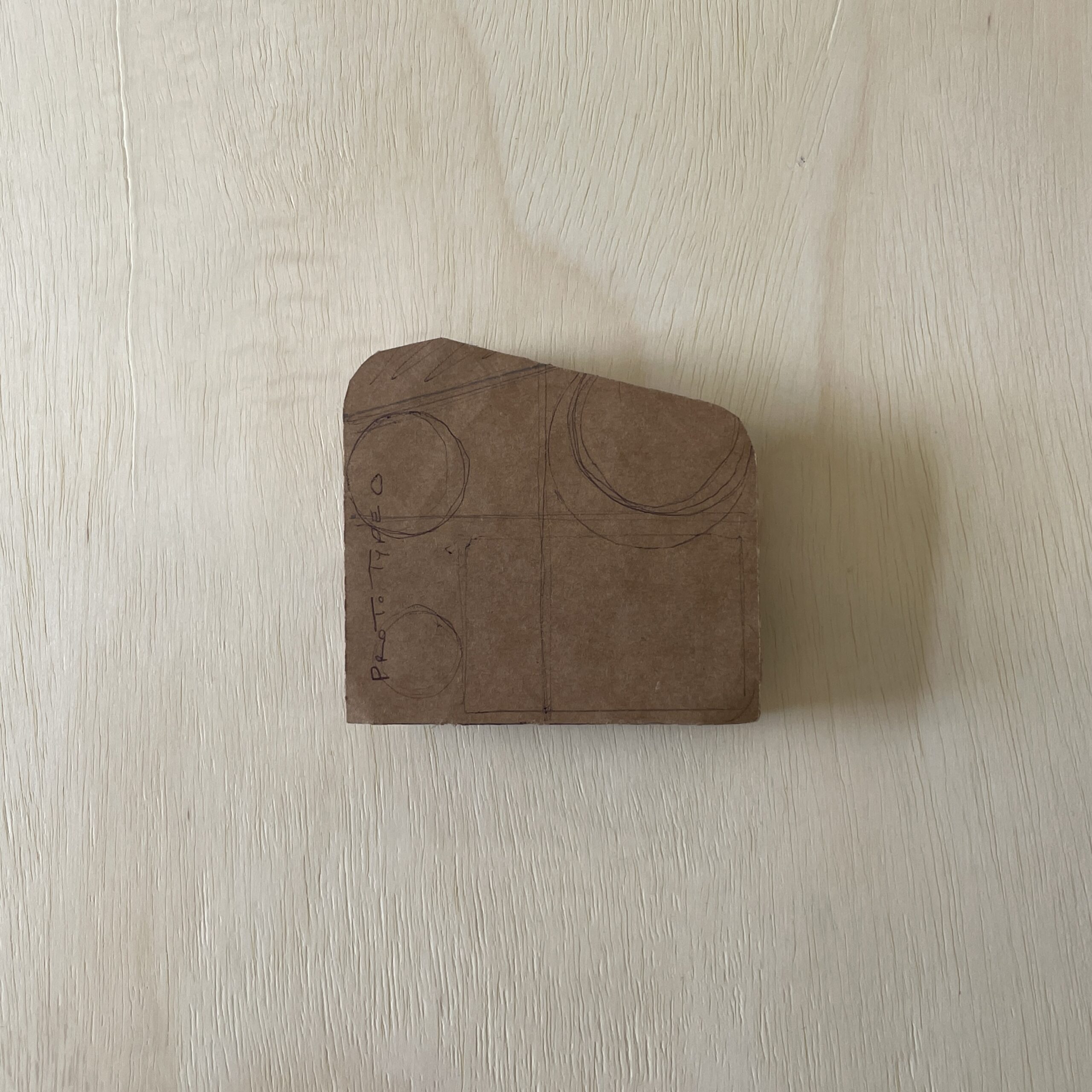 A piece of brown cardboard with a rounded corner features hand-drawn geometric shapes and text labeled "Proto Type 0." It is positioned on a light wooden surface with a visible grain pattern. The background is light-colored and slightly textured.