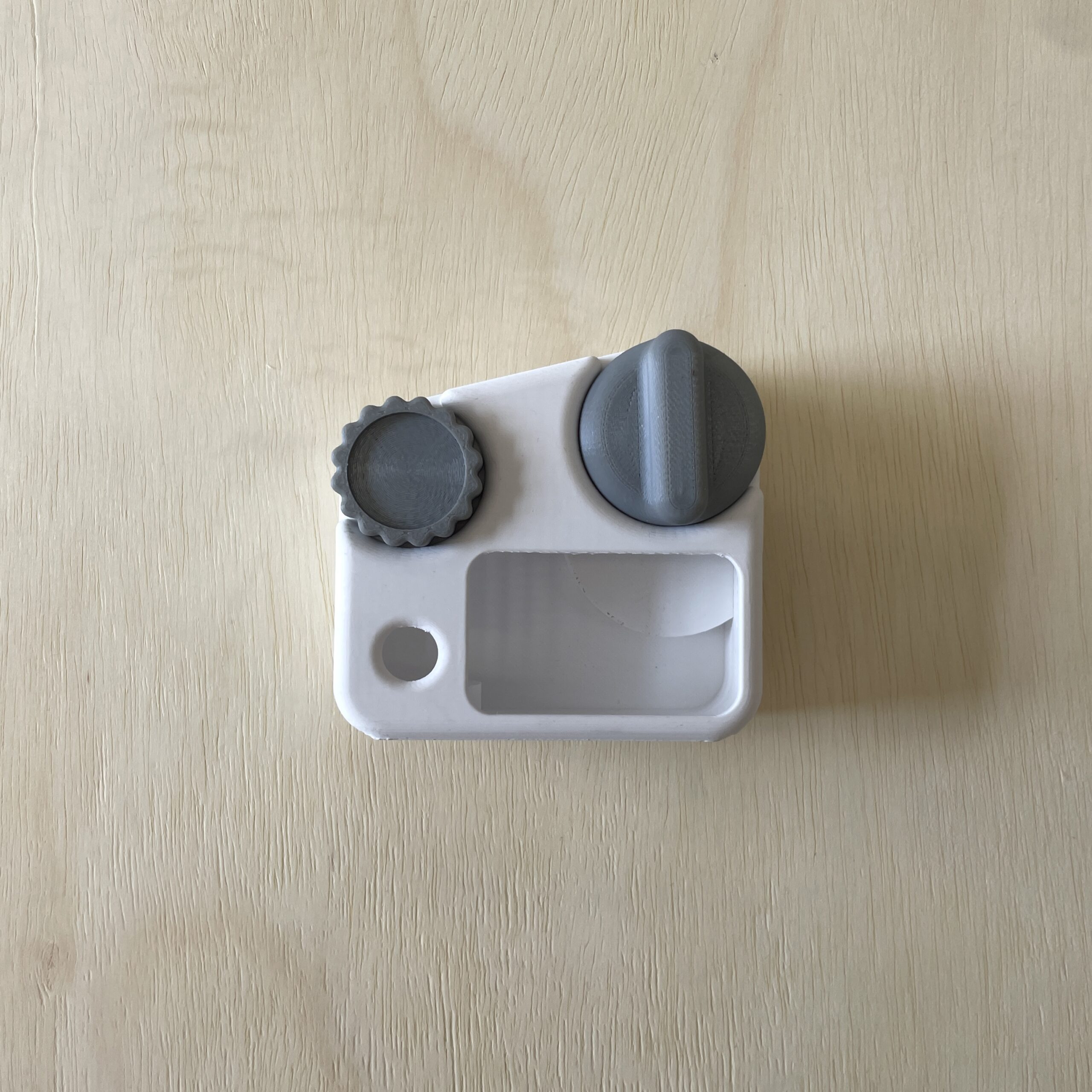 A small white plastic object with two gray knobs. The left knob is round with ridges, and the right knob is a rounded rectangle. The front features a small rectangular opening and a circular hole. The object is placed on a light wood surface.