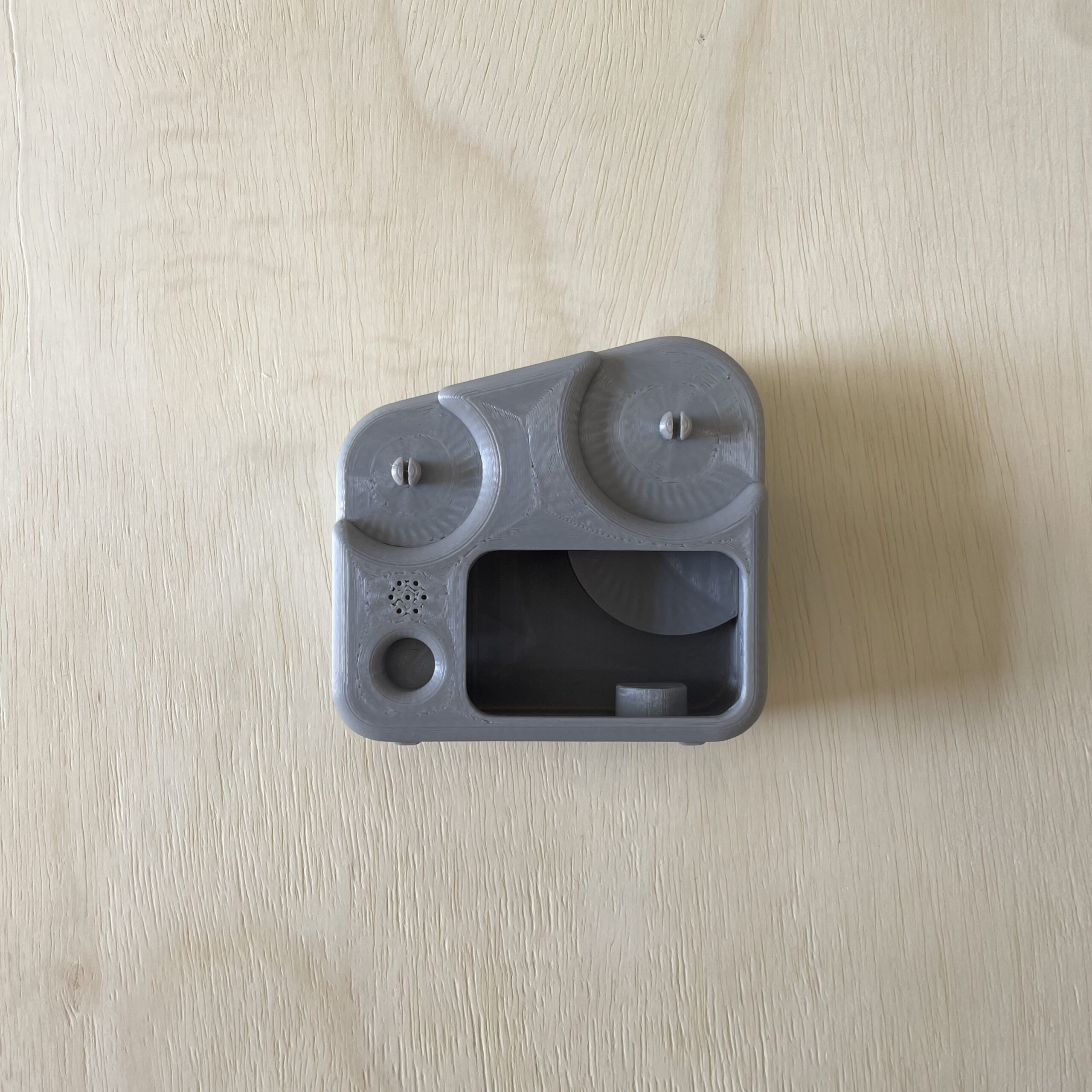A gray, rectangular 3D-printed object resembling a compact device with two circular dials on the top. The front features an open rectangular slot and additional smaller holes. The texture is smooth with visible print lines, sitting on a light wooden surface.
