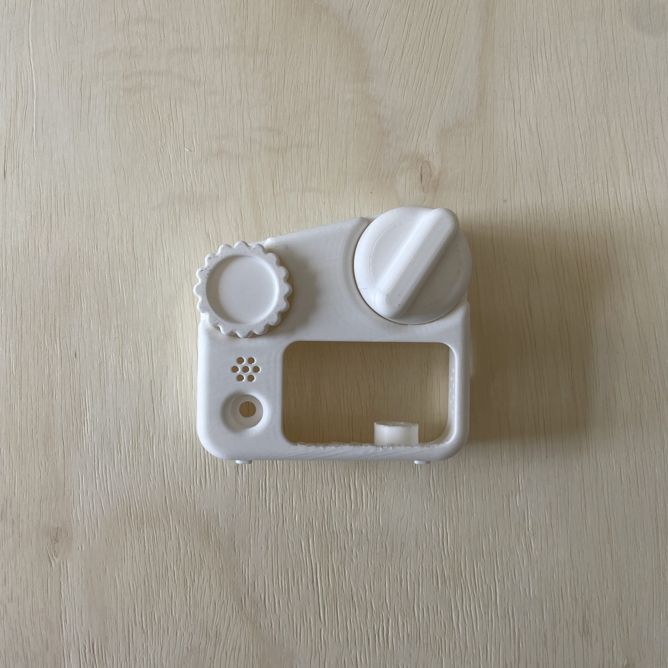 A white plastic component resembling a camera body is displayed on a light wooden surface. It features a large round knob on the right, a smaller gear-like knob on the left, and a rectangular opening in the center with a small button below it.