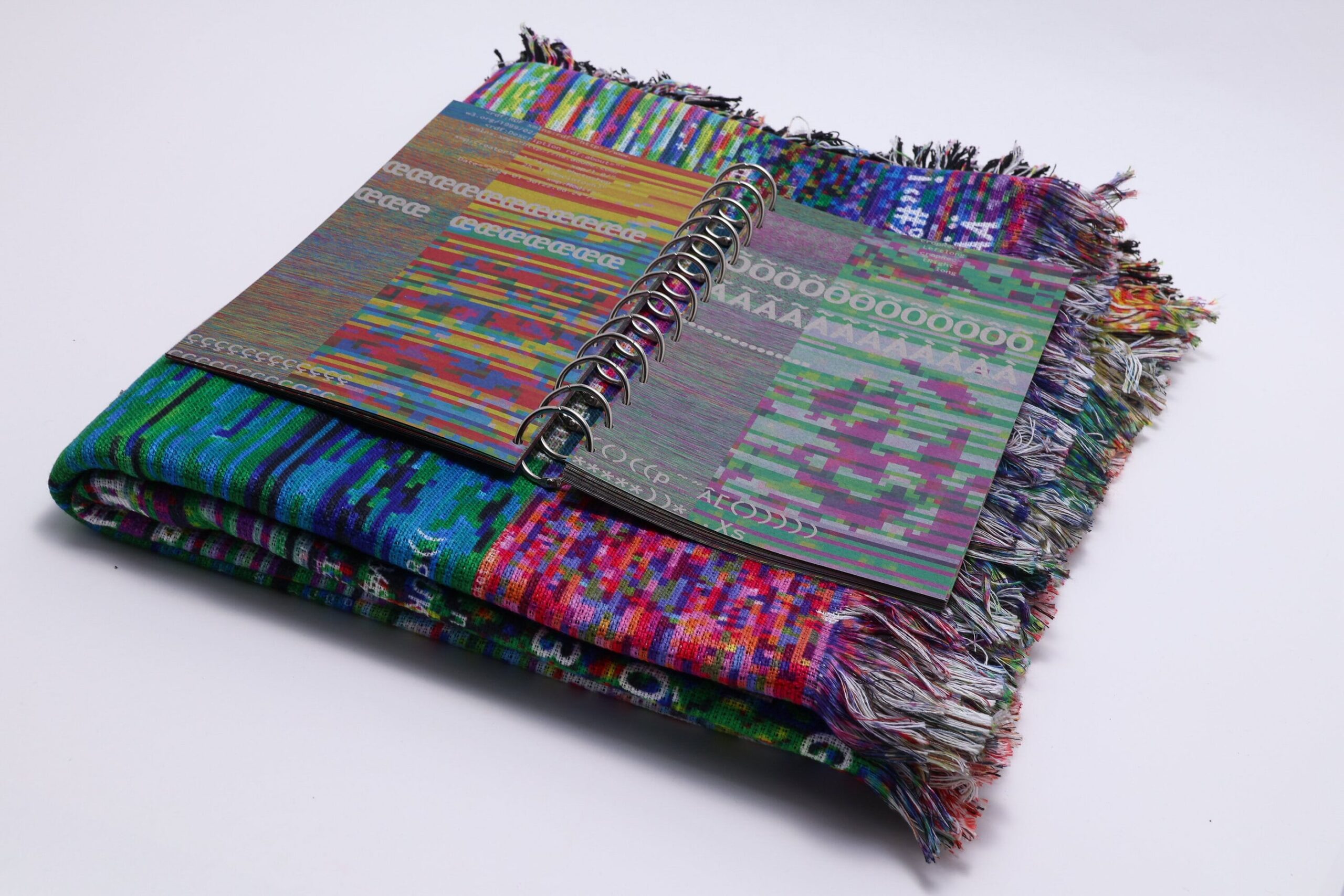 An open notebook with a spiral binding rests atop a colorful woven blanket. The notebook features vibrant patterns in various colors and shapes, while the blanket showcases intricate designs primarily in green, purple, and blue hues, along with textured fringes. The arrangement presents a fusion of artistic designs and textiles.