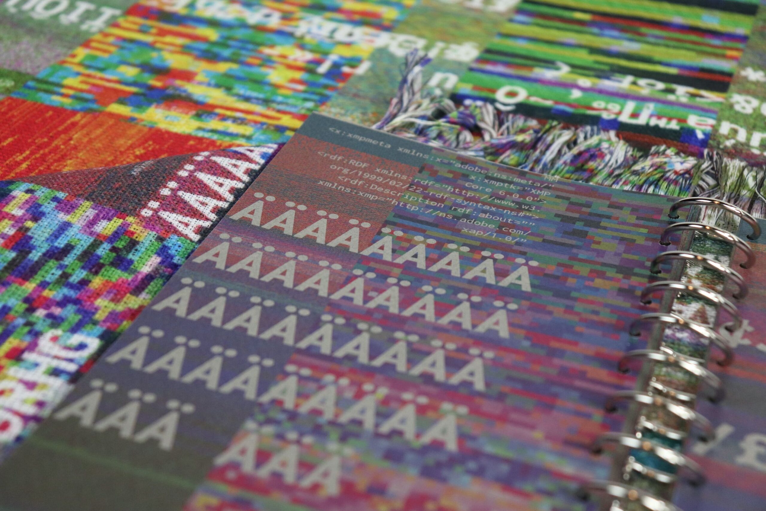 A close-up image of a colorful notebook featuring a variety of textured pages with vibrant patterns. Some pages display the letter "A" with diacritics in different styles and colors, while the background has a mix of abstract, pixelated designs. The metal rings of the notebook are visible on the right side, and a portion of the spine shows printed text, adding to the artistic composition.