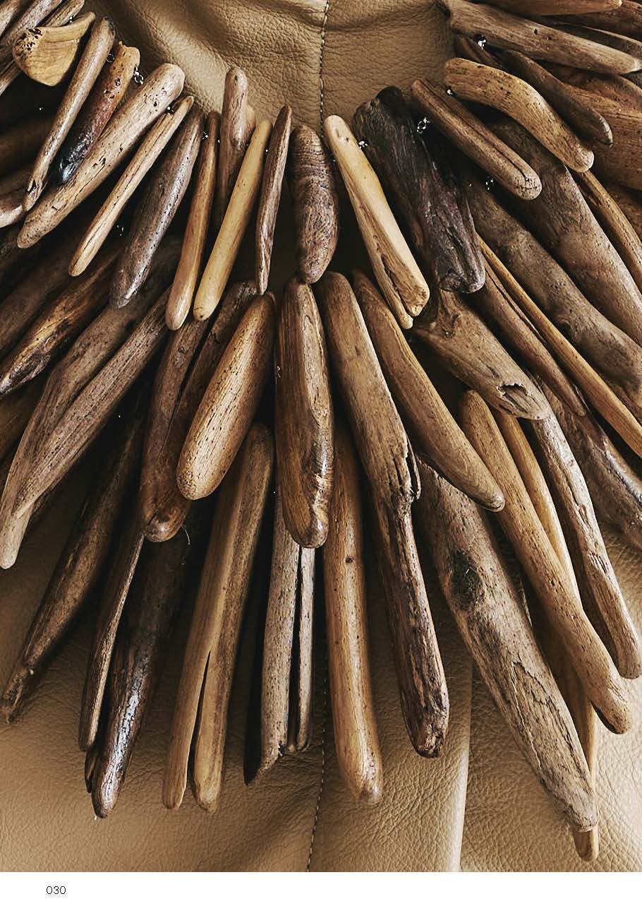 Close-up of a unique necklace made of elongated pieces of driftwood, arranged in a fan shape. The wooden pieces vary in size and shade, creating a textured and organic look. The background shows a subtle leather surface.