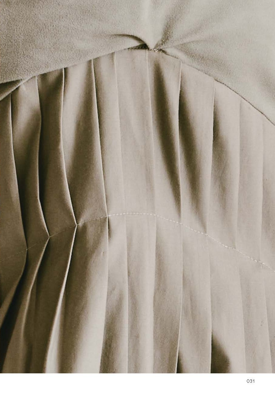 Close-up image of a fabric with soft, pleated textures in neutral tones. The fabric is divided into sections, featuring vertical pleats that create a flowing appearance. The top portion appears smooth and slightly gathered. A subtle seam runs horizontally across the pleats, enhancing the detailed craftsmanship of the garment.