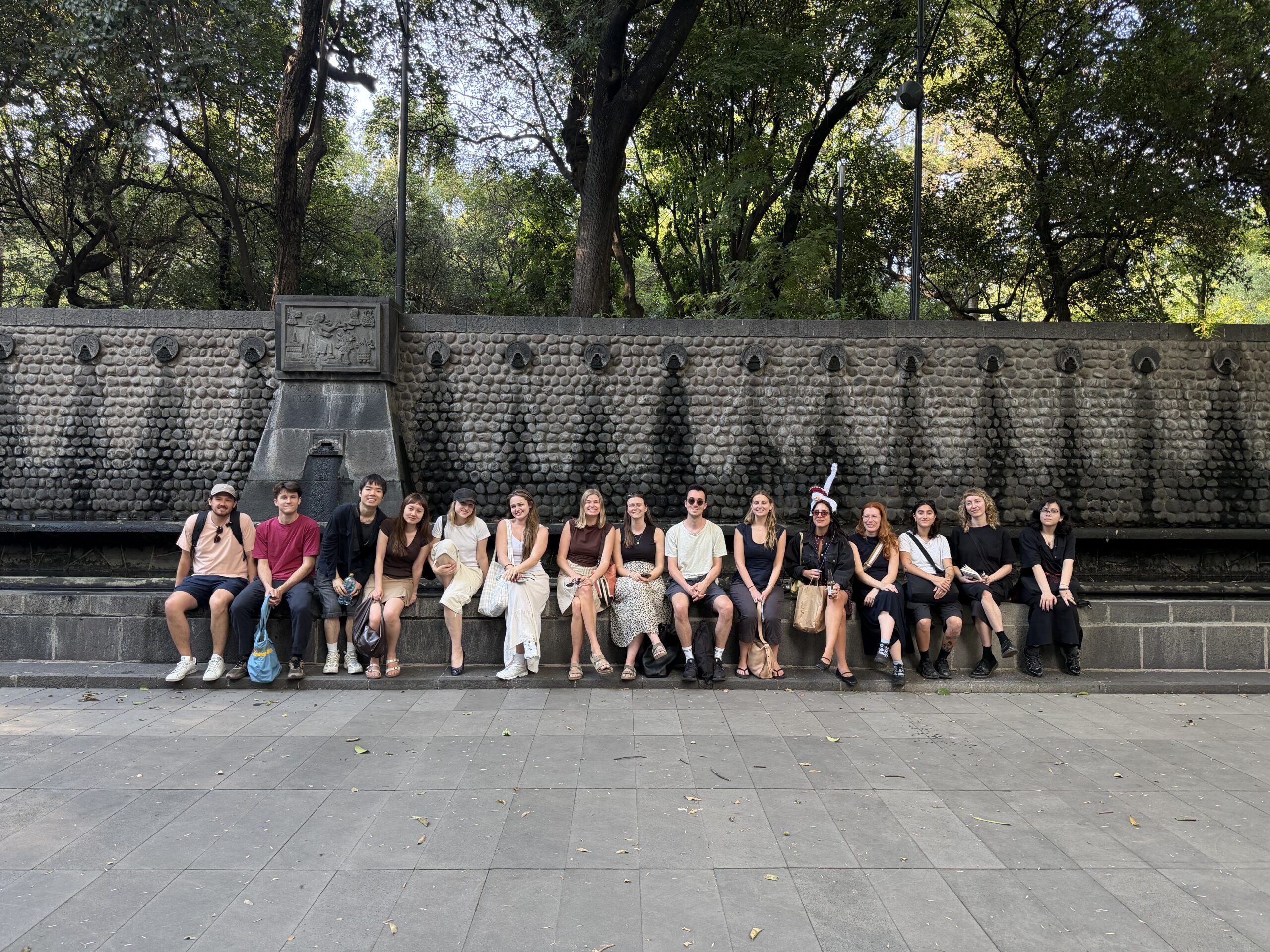 A group of 14 people are sitting casually on a stone bench in a park. They are arranged in two rows against a stone wall with decorative patterns. The setting is surrounded by trees, and there are some fallen leaves on the ground. The group appears to be enjoying a sunny day, wearing a mix of casual clothing in various colors.