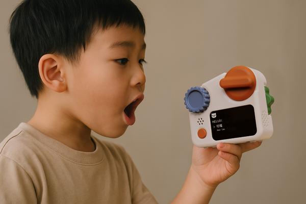 Two small electronic devices are displayed on a gray background. Each has a white body with colorful knobs: one is orange, and the other is blue. They feature screens with text in both English and Chinese, indicating conversational prompts or greetings. One device shows "HELLO! HOW ARE YOU?" while the other displays, "I AM GOOD! TODAY I WENT TO THE ZOO!" Each has a speaker icon and additional buttons on the side.