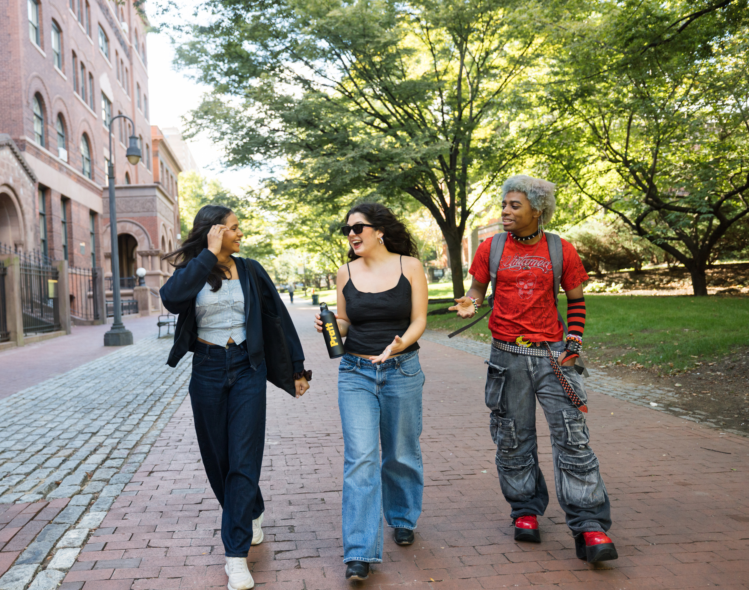 Three young adults are walking together on a brick path surrounded by trees. Two women, one in a cropped striped top and wide-leg jeans, and another in a black tank top and blue jeans, are smiling and conversing. The third person, a man with dyed hair and a red shirt, is wearing cargo pants and platform shoes, engaging with the women. The background features a brick building and greenery.