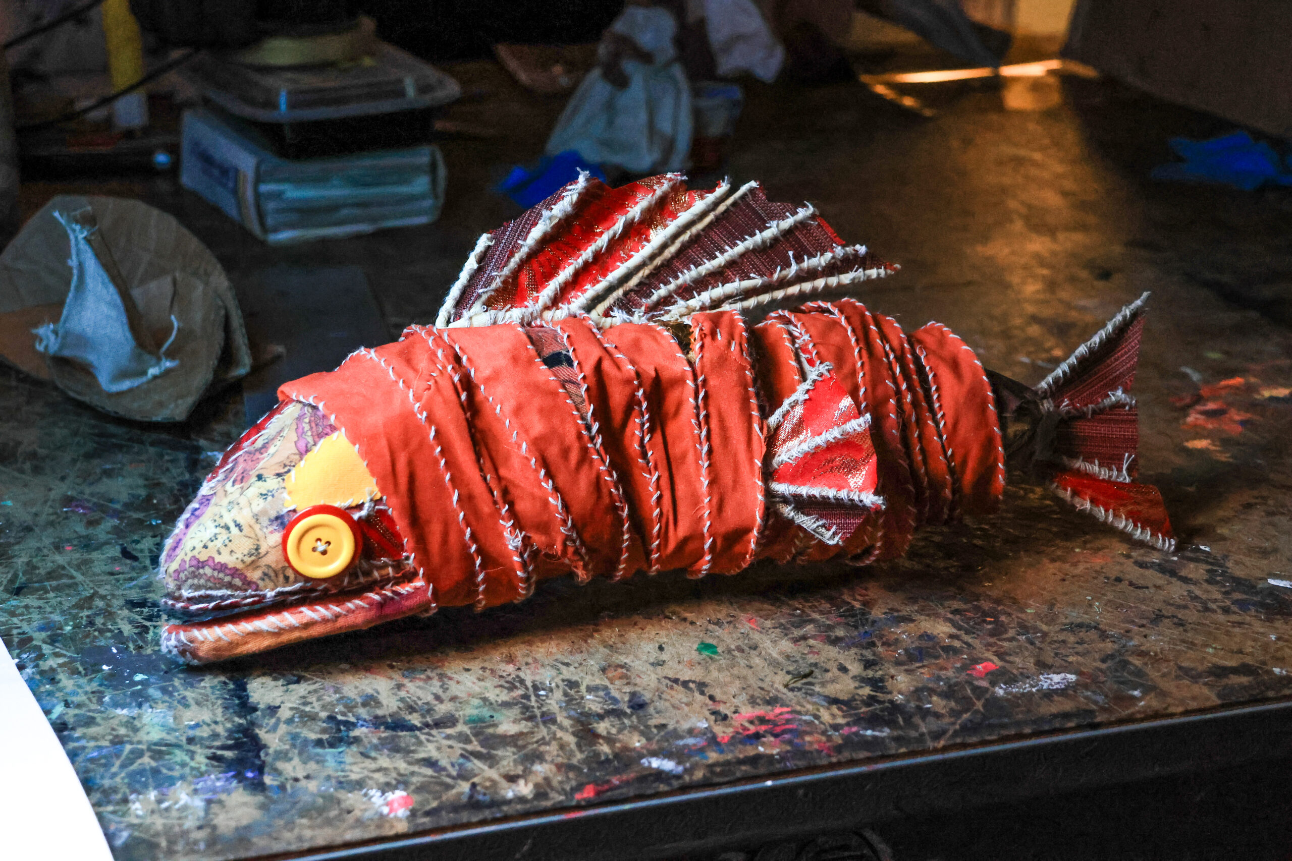 A creatively crafted fish sculpture made from various textiles, primarily red fabric, is placed on a cluttered wooden table. The fish has decorative scales and a yellow button for an eye, showcasing a unique and artistic design. Pieces of cardboard and other materials are partially visible in the background, adding to the eclectic workspace ambiance.