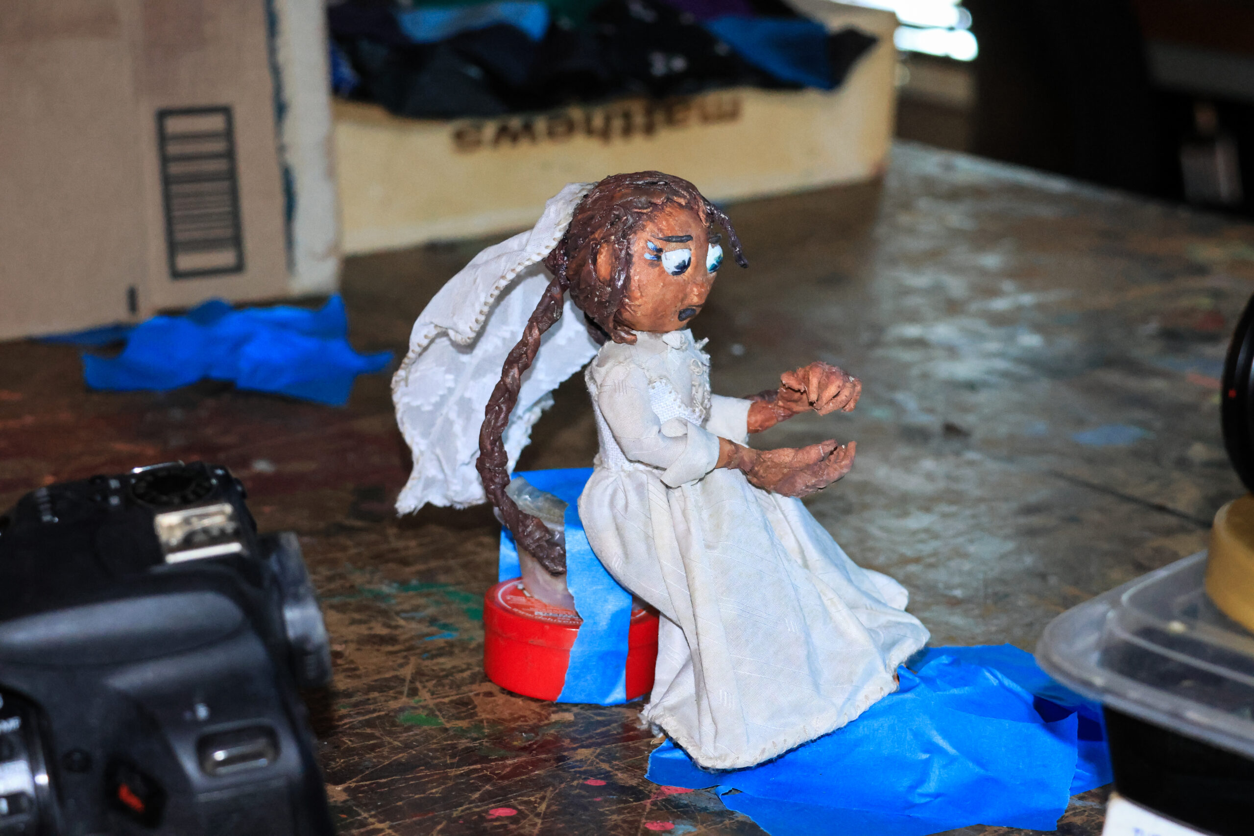 A handmade doll sits on a small, circular red base, wearing a white dress and veil. The doll has braided hair and appears to be gesturing with its hands. Surrounding it are blue painter's tape and various objects including a camera and a cardboard box in the background, with scattered items on the table's surface. The lighting is warm, highlighting the details of the doll.