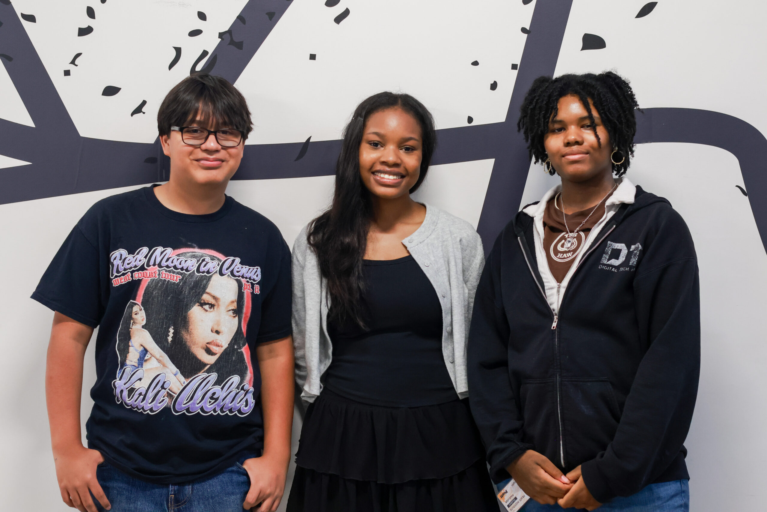 Three young individuals are standing together in front of a light colored wall with abstract black designs. The person on the left is wearing a black t-shirt with a graphic print and glasses. The middle person is smiling and wearing a gray cardigan over a black top and a skirt. The person on the right is in a dark hoodie with a white lining and is also smiling.