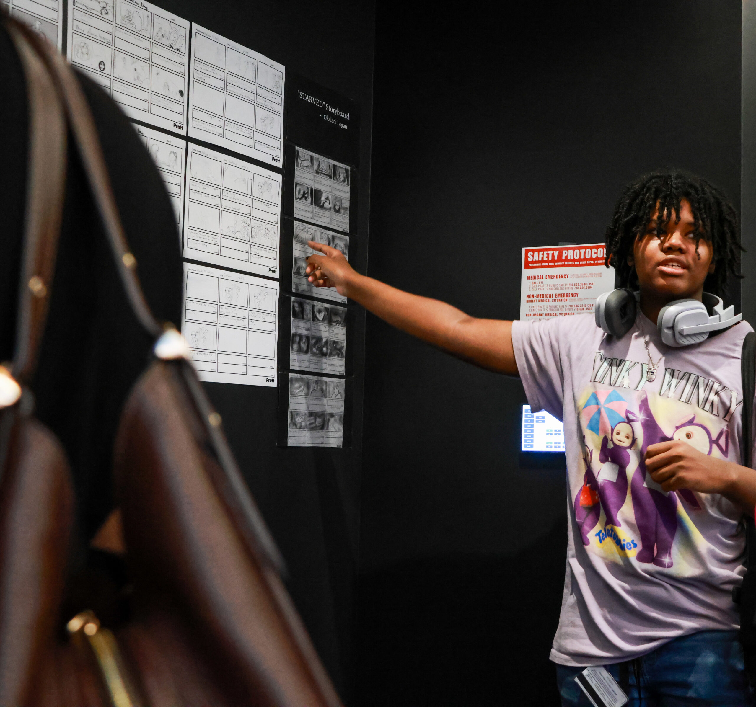 A young person with shoulder-length curly hair is pointing at a storyboard mounted on a wall. They are wearing headphones and a t-shirt with cartoon characters. In the background, there is a warning sign regarding safety protocols. A part of a brown bag is visible on the left side of the image.