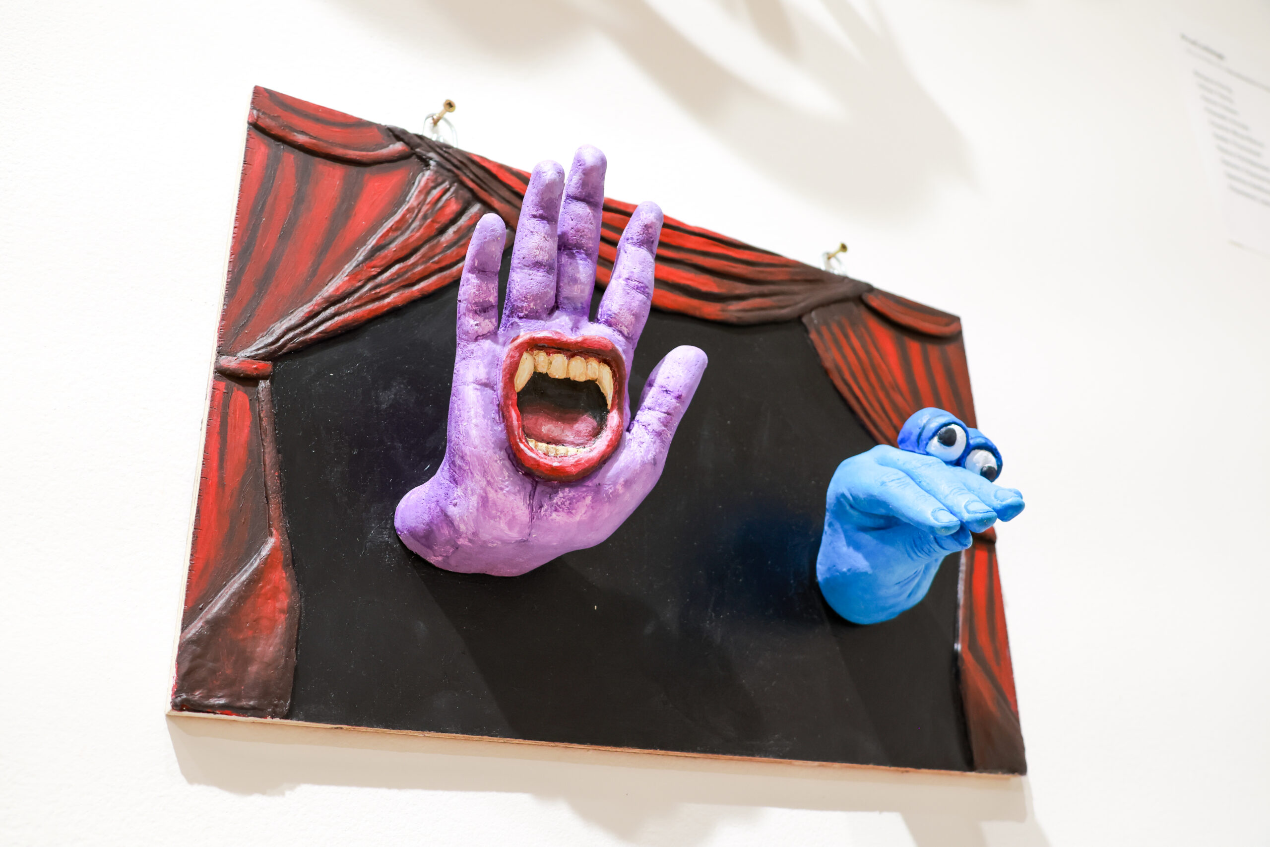 A colorful mixed-media artwork featuring two hands protruding from a black background framed by red curtains. The left hand is purple with an open mouth showing teeth, while the right hand is blue with a pair of cartoonish eyes. The piece combines vibrant colors and playful elements, creating an engaging visual effect.
