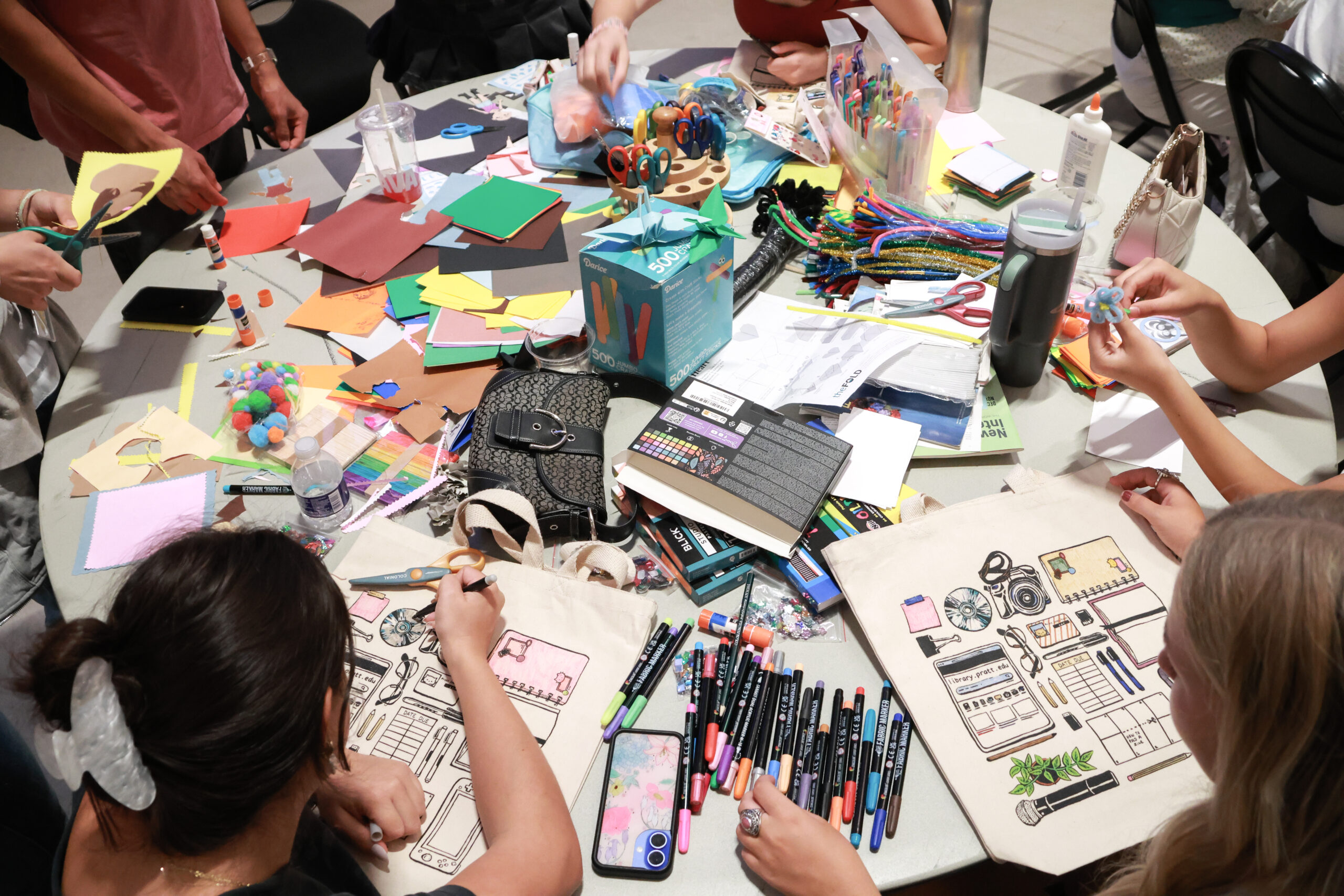 A cluttered table filled with various crafting supplies, including colored paper, scissors, glue, markers, and pom-poms. Several people are engaged in a creative activity, with one person using scissors and another drawing on a tote bag. The scene captures an interactive and artistic atmosphere, with supplies scattered around and vibrant colors present.