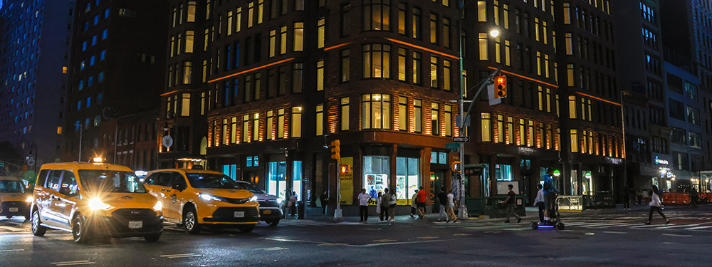 T bustling intersection of 14th Street and 6th Avenue in Manhattan at night, featuring two yellow taxis and a minivan in the foreground.
