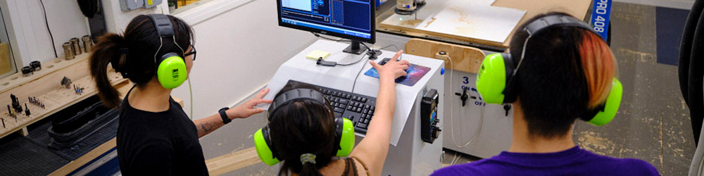 A group of three individuals is engaged in a collaborative project in a workshop. They are wearing bright green headphones and focused on a computer screen displaying software.