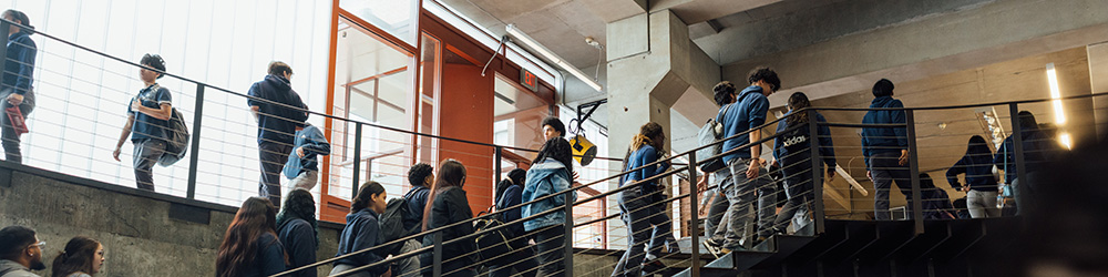 A busy staircase inside a modern building, with students in casual clothing walking up and down. Some students are carrying backpacks. Large windows allow natural light to illuminate the space, and there are metal railings and concrete walls. The atmosphere appears lively and dynamic.