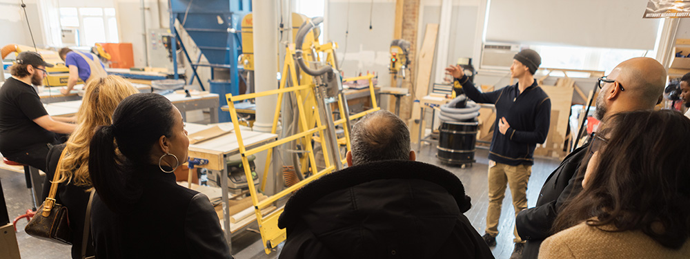 A group of people watches a presentation in a workshop. A man in a dark sweater gestures while speaking, surrounded by machinery and workstations.