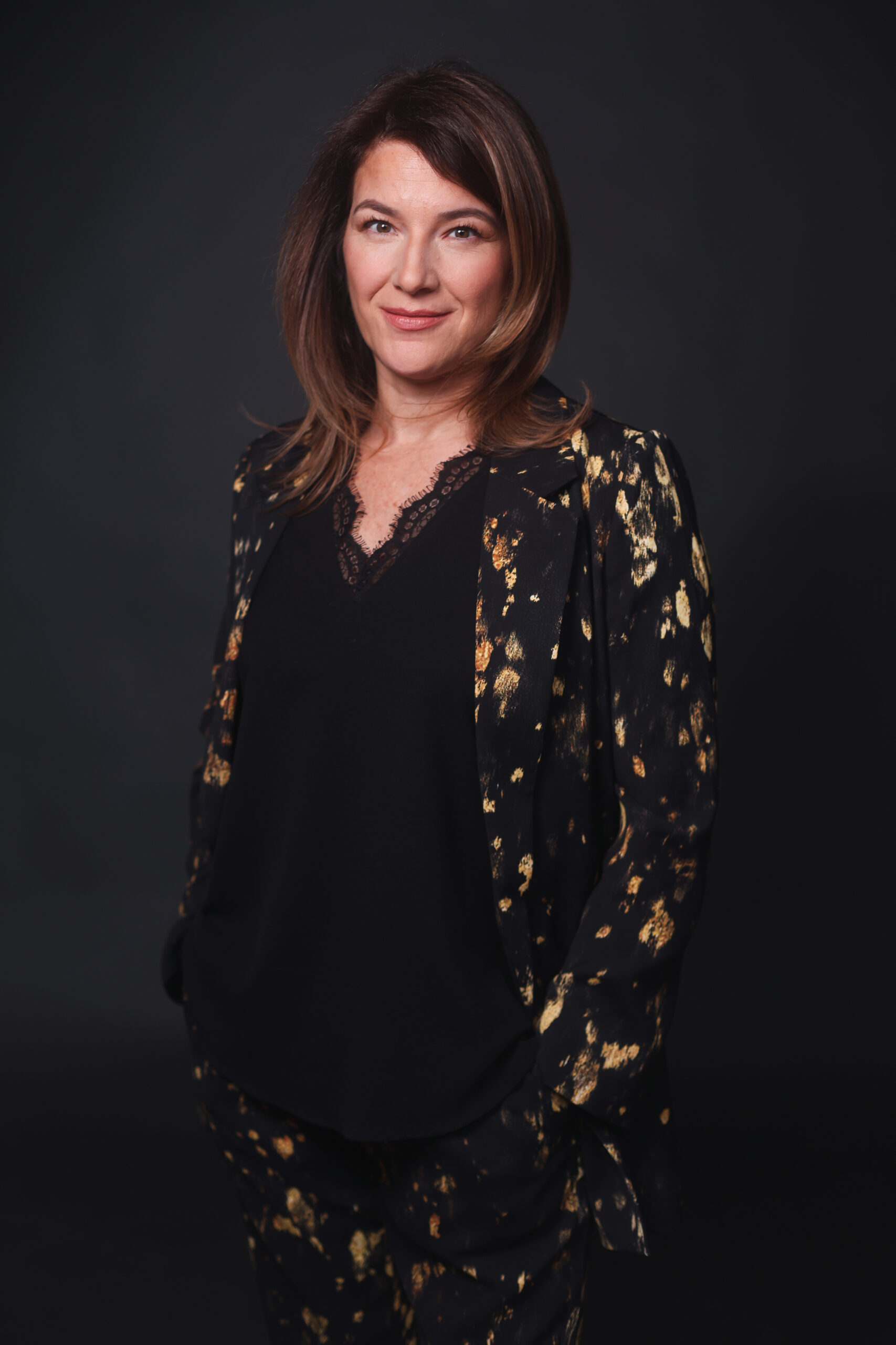 Studio portrait of a person standing against a dark background, wearing a black outfit with a black jacket and matching pants featuring a metallic gold abstract pattern. They face the camera with a slight smile, hands in their pockets, under soft studio lighting.