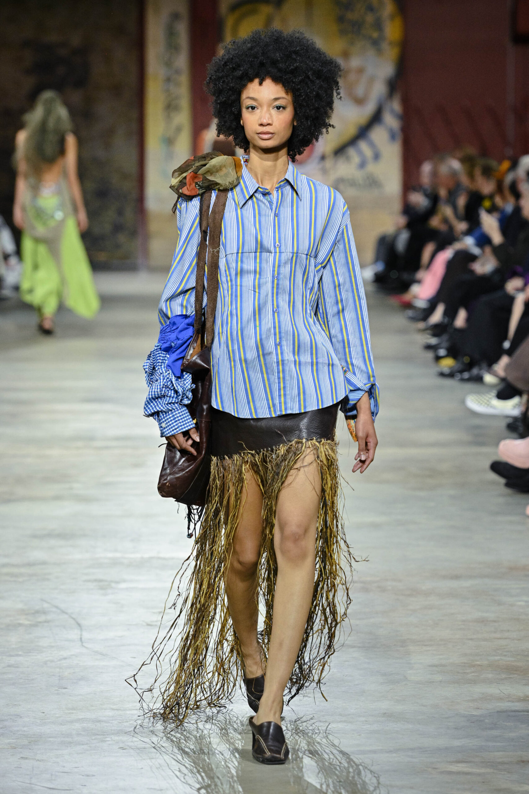 A model walks down a runway wearing a striped oversized shirt with billowy sleeves, a brown leather skirt embellished with fringe, and black shoes. She carries a brown bag with a colorful patch. In the background, another model is seen in a flowing green outfit, while an audience observes along the runway. The setting features a modern, artistic atmosphere.