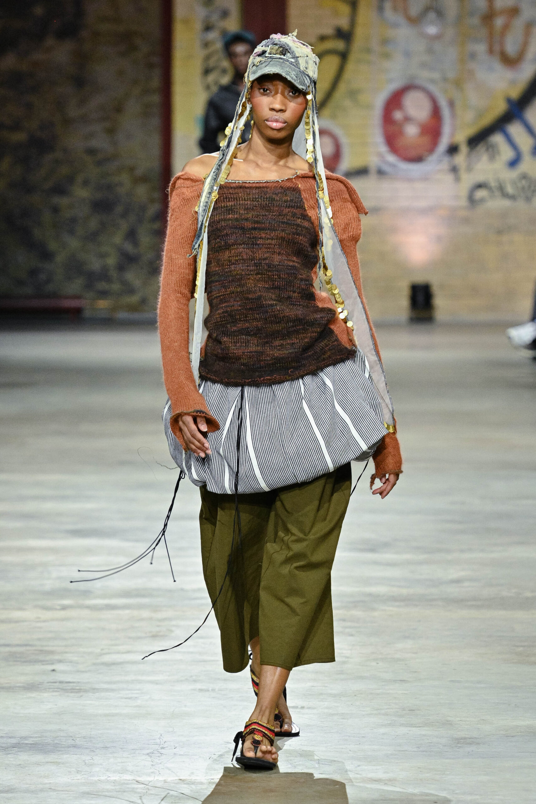 A model walks down a runway wearing a unique outfit that combines a brown off-the-shoulder knit top with a layered striped skirt and olive green pants. The ensemble is accessorized with a denim cap featuring decorative yarn strands. The background features a textured surface and graffiti-like artwork, emphasizing a vibrant fashion show atmosphere.
