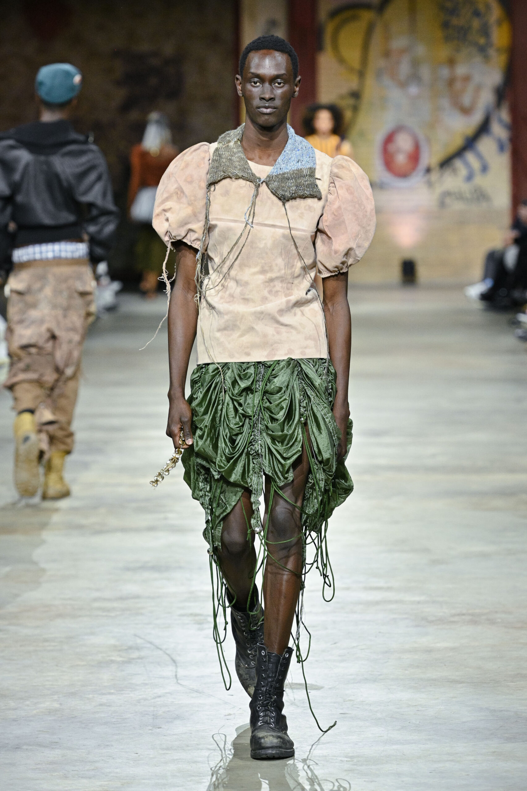 A model walks down a runway wearing an avant-garde outfit featuring a puffed-sleeve beige top and a green, draped skirt with textured detailing. The model's look is completed with black combat boots. In the background, additional models are seen in various outfits, set against a backdrop of artistic murals.