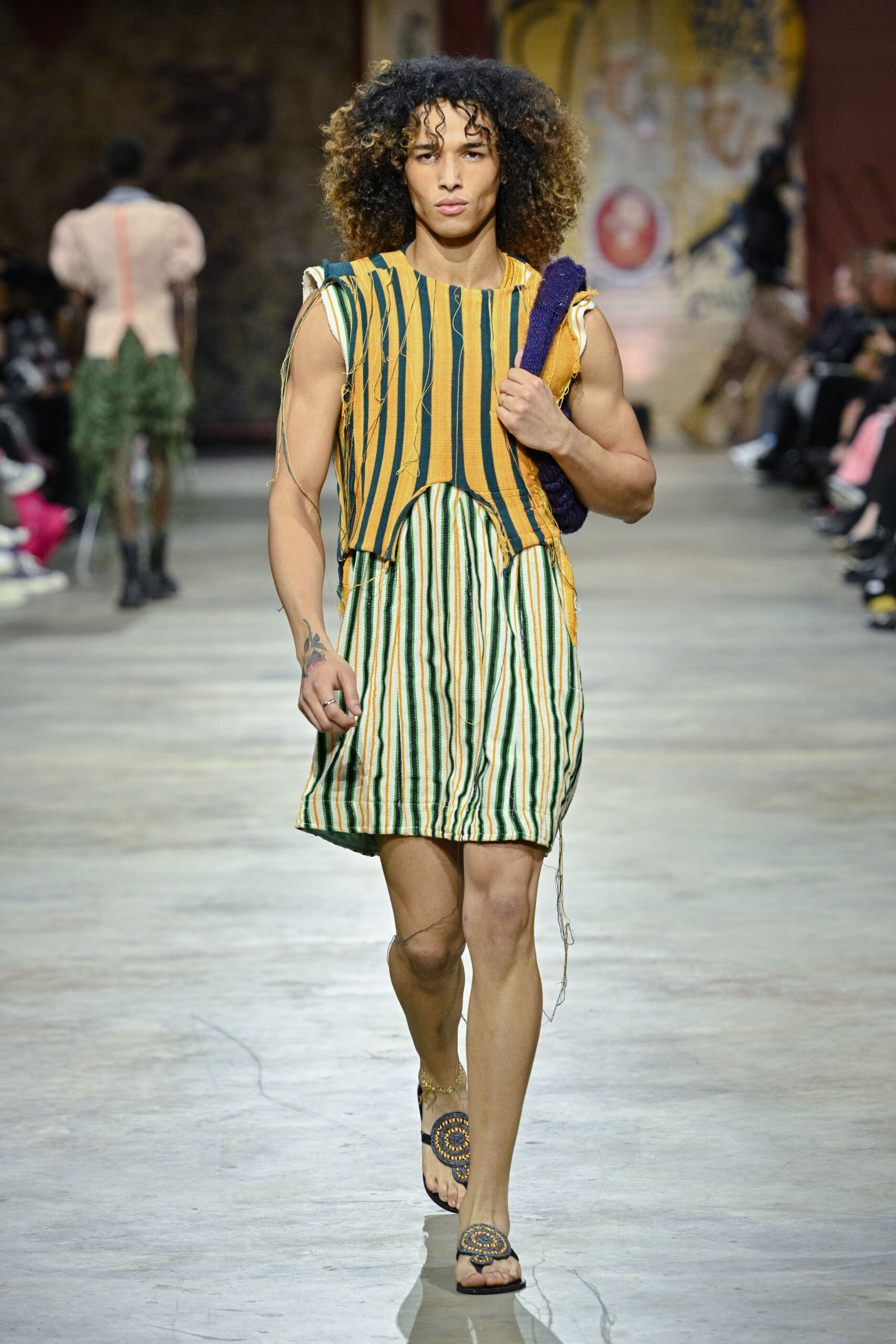 A male model walks down a runway wearing a colorful, vertically striped dress with a loose fit, showcasing a blend of yellow, green, and orange hues. He carries a textured bag and wears decorative sandals. The backdrop features an audience and artistic wall designs, adding to the fashion show atmosphere.