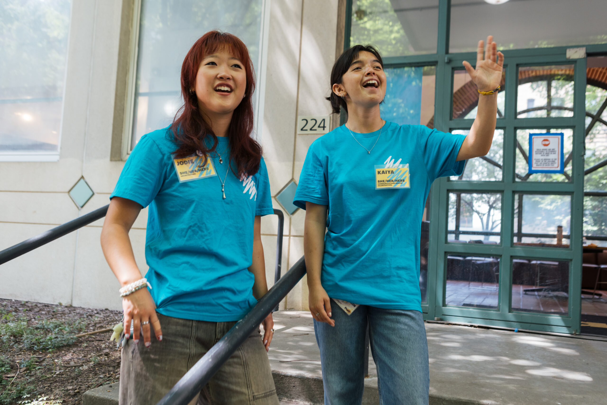 Two young people, both wearing bright blue t-shirts, stand at the entrance of a building. One has red hair and is smiling as they gesture with their hand, while the other has dark hair and is laughing. They appear to be in a welcoming or enthusiastic moment, with sunlight filtering through nearby trees. A sign is visible on the door behind them.