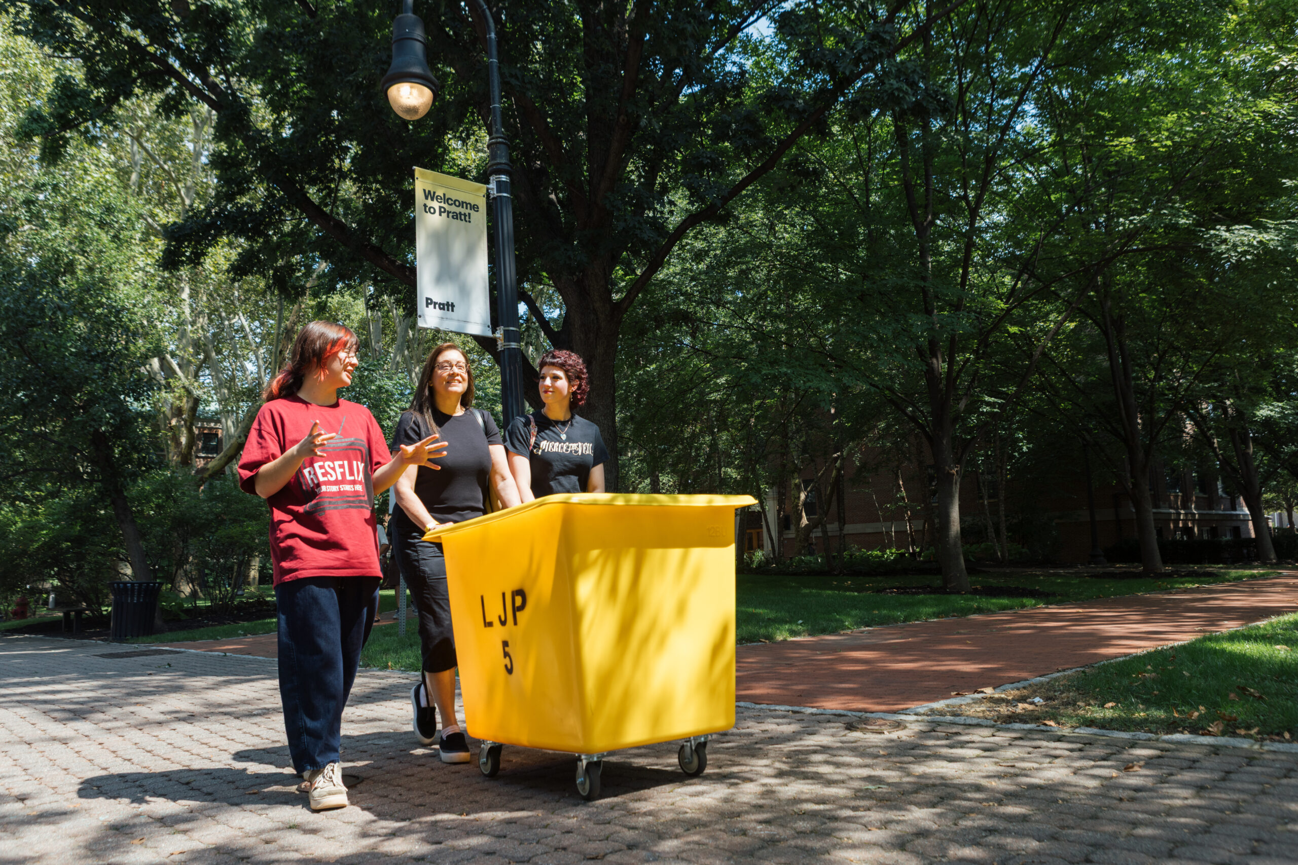 Three individuals are standing near a yellow cart in a park-like setting, surrounded by trees. One person, wearing a red shirt, appears to be speaking animatedly, while another person, dressed in black, is listening attentively. A banner hanging from a lamp post reads "Welcome to Pratt!" The ground is made of cobblestones, and there's a sidewalk visible in the background.